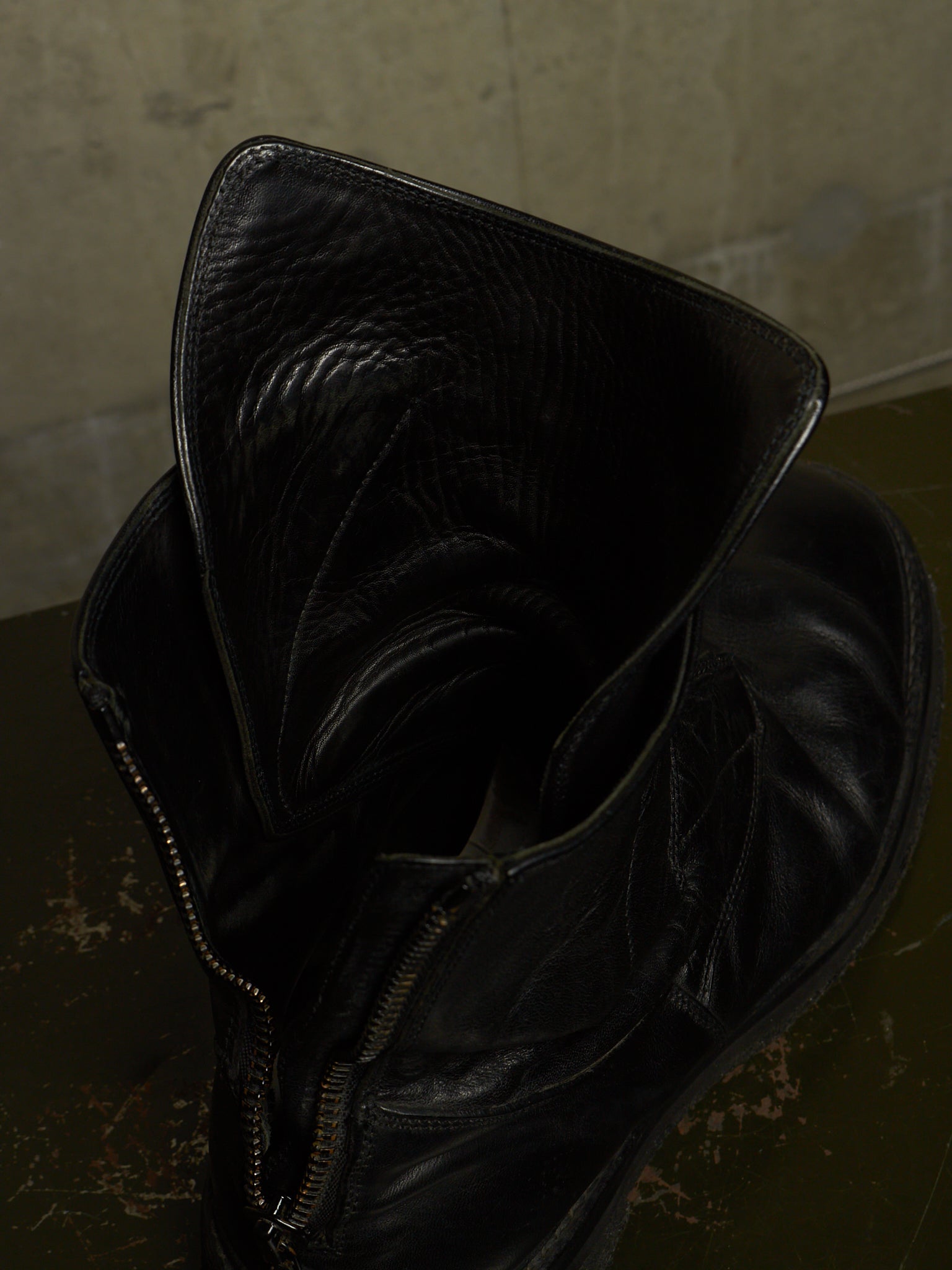 A close-up of a black leather ankle boot with a wide, folded-up collar and a side zipper, resting on a dark textured surface against a neutral concrete wall.