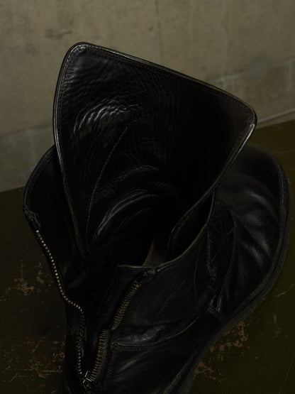 A close-up of a black leather ankle boot with a wide, folded-up collar and a side zipper, resting on a dark textured surface against a neutral concrete wall.