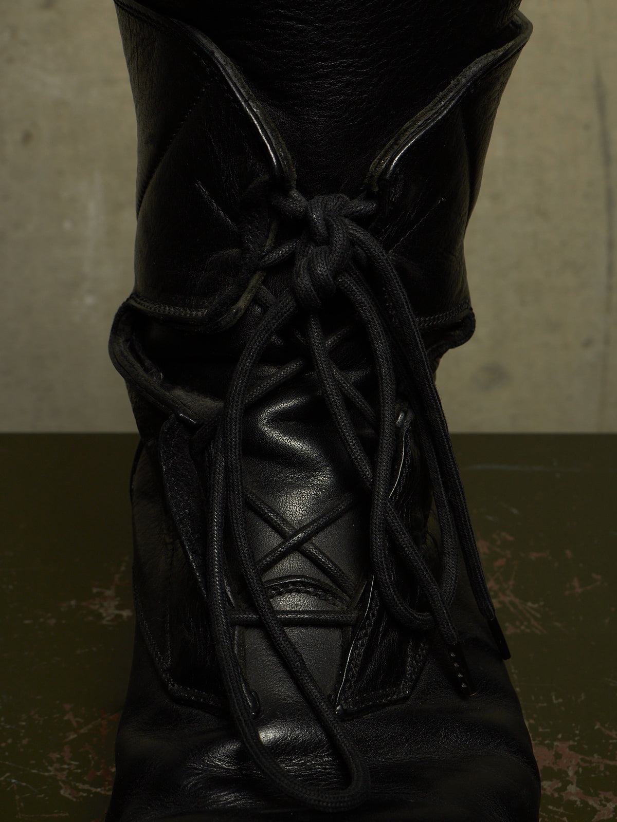 A close-up of a black leather lace-up boot with thick black laces, set against a dark olive-green surface and a textured gray background.