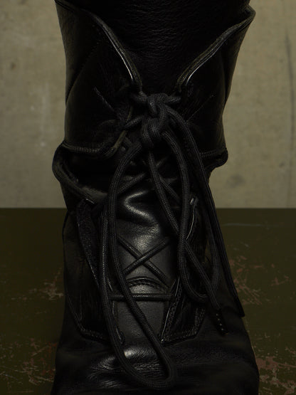 A close-up of a black leather lace-up boot with thick black laces, set against a dark olive-green surface and a textured gray background.