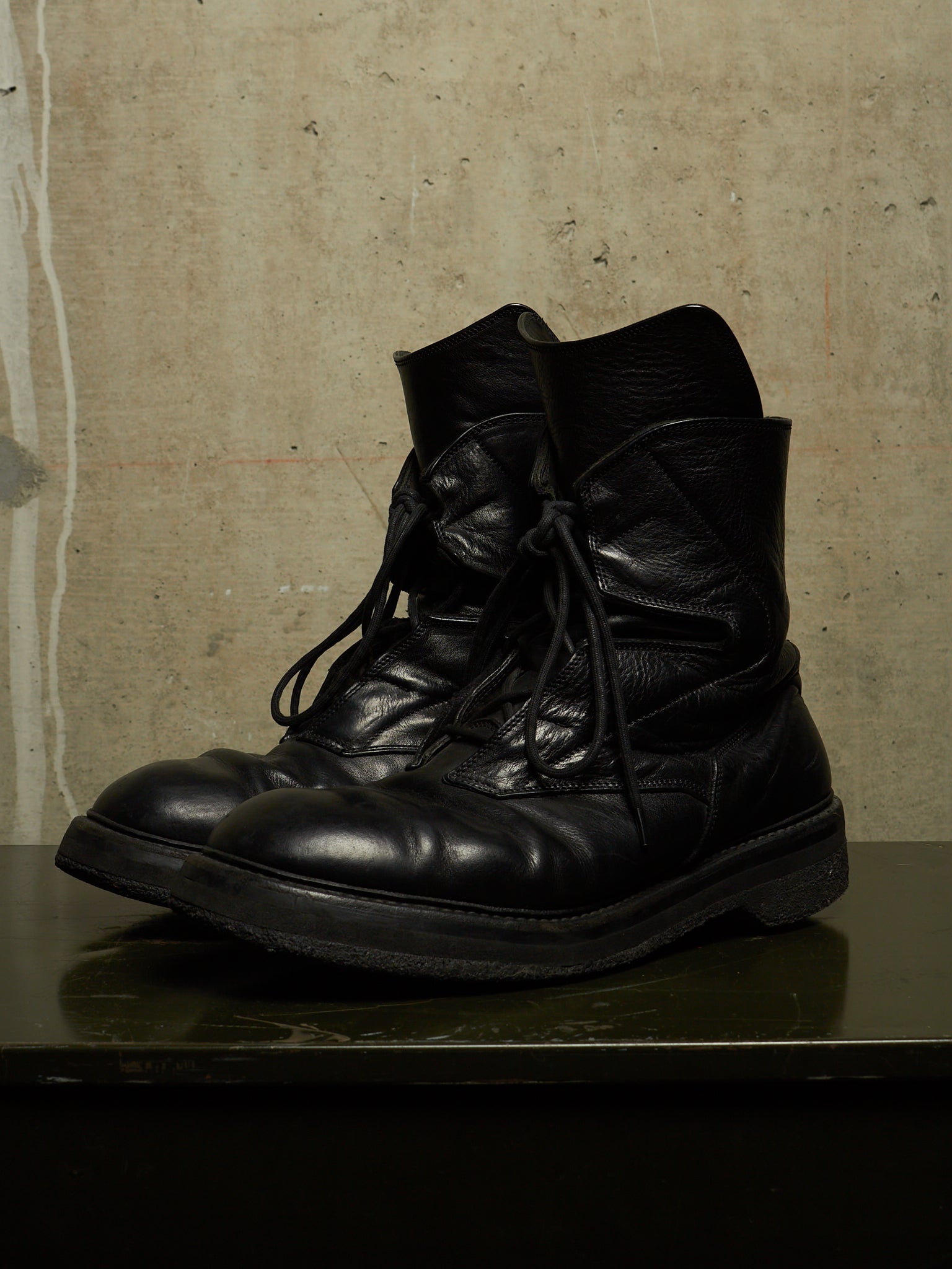 A pair of black leather lace-up combat boots with thick soles, displayed on a dark reflective surface against a textured beige wall.