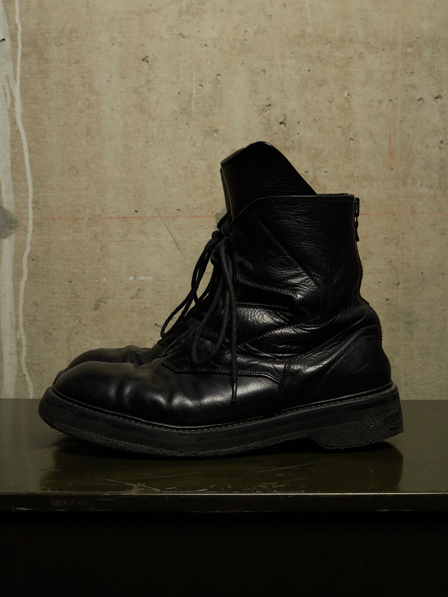 A pair of black leather lace-up ankle boots with a rugged sole, resting on a dark reflective surface against a textured concrete wall.