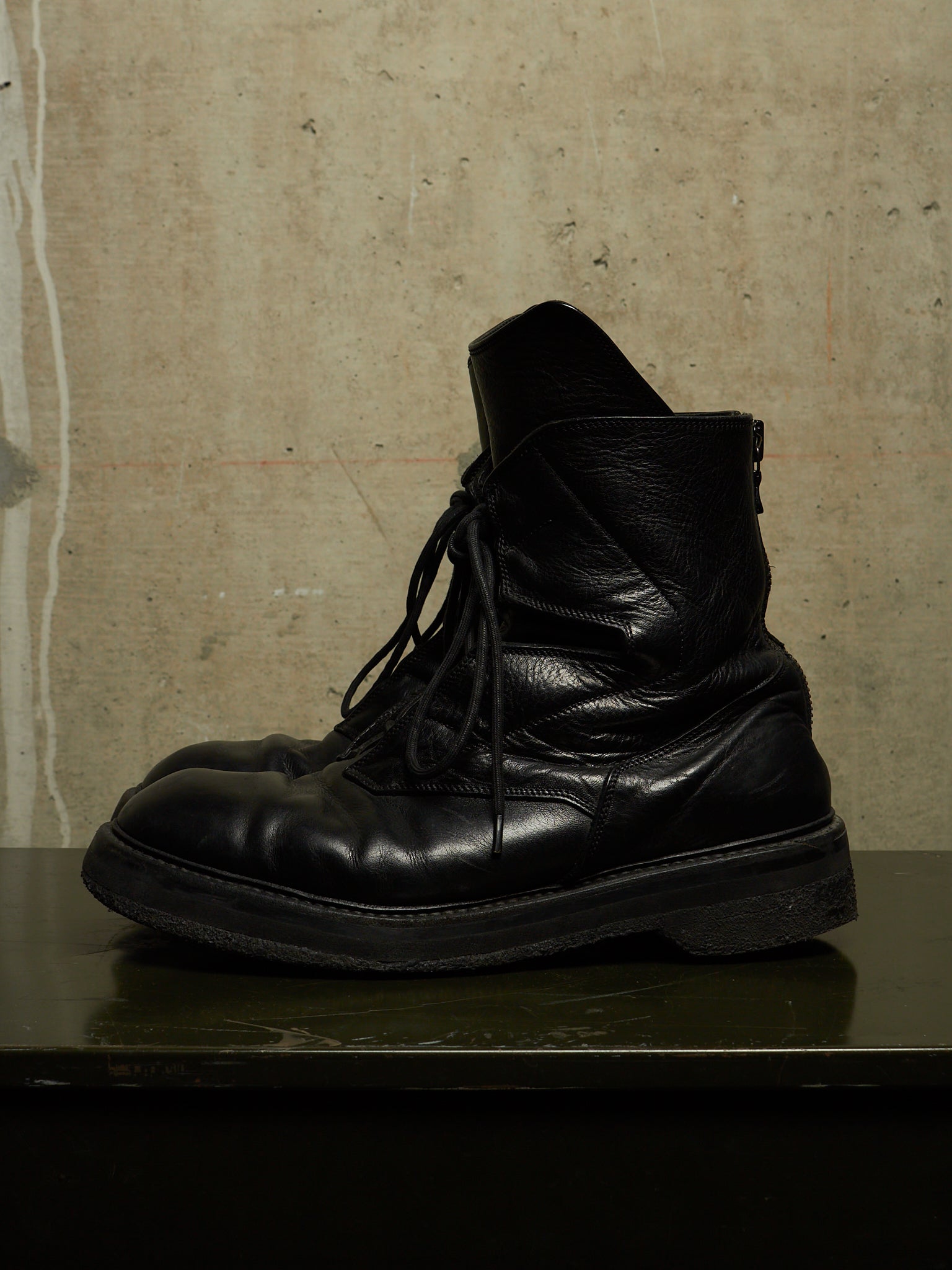 A pair of black leather lace-up ankle boots with a rugged sole, resting on a dark reflective surface against a textured concrete wall.
