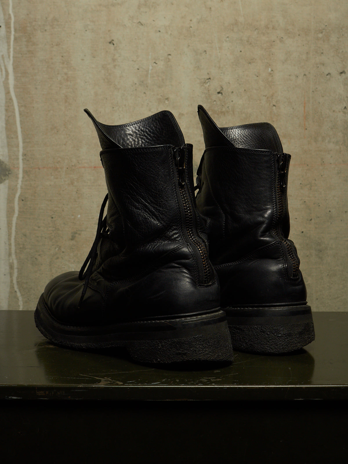 A pair of black leather lace-up boots with side zippers, shown from the back on a dark surface against a textured concrete wall.