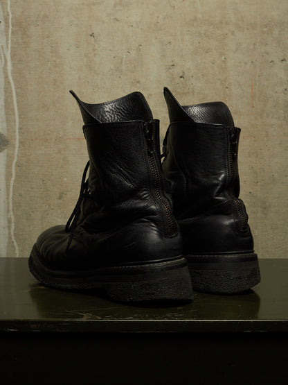 A pair of black leather lace-up boots with side zippers, shown from the back on a dark surface against a textured concrete wall.