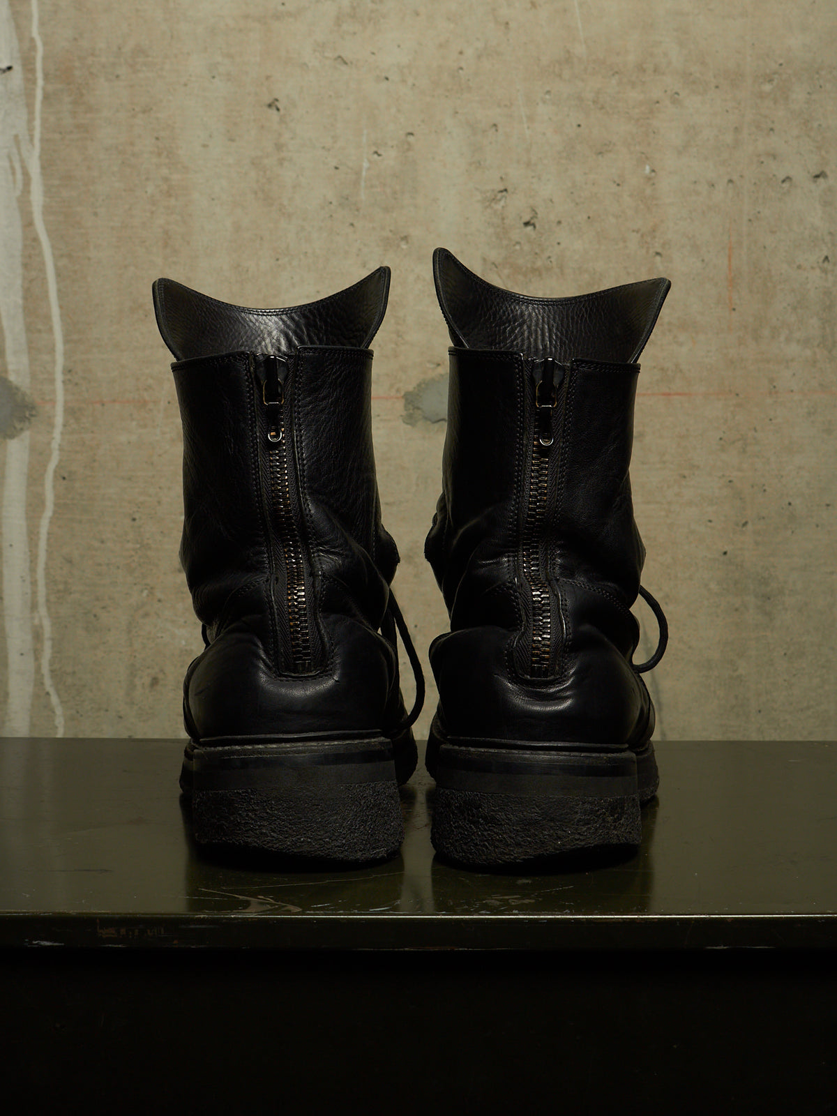A pair of black leather ankle boots with side zippers, shown from the back on a dark reflective surface against a textured concrete wall.