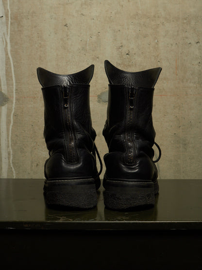 A pair of black leather ankle boots with side zippers, shown from the back on a dark reflective surface against a textured concrete wall.