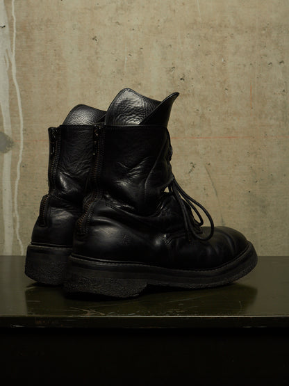 A pair of black leather lace-up ankle boots with side zippers, displayed on a dark reflective surface against a textured concrete wall.