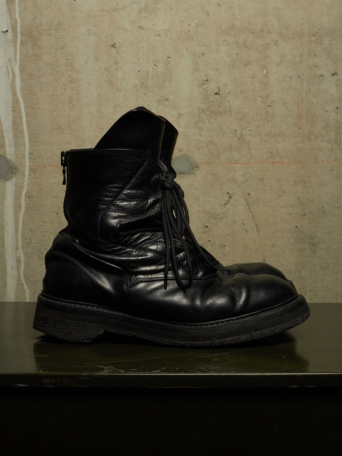 A single black leather lace-up ankle boot with a side zipper, resting on a dark reflective surface against a textured concrete wall.
