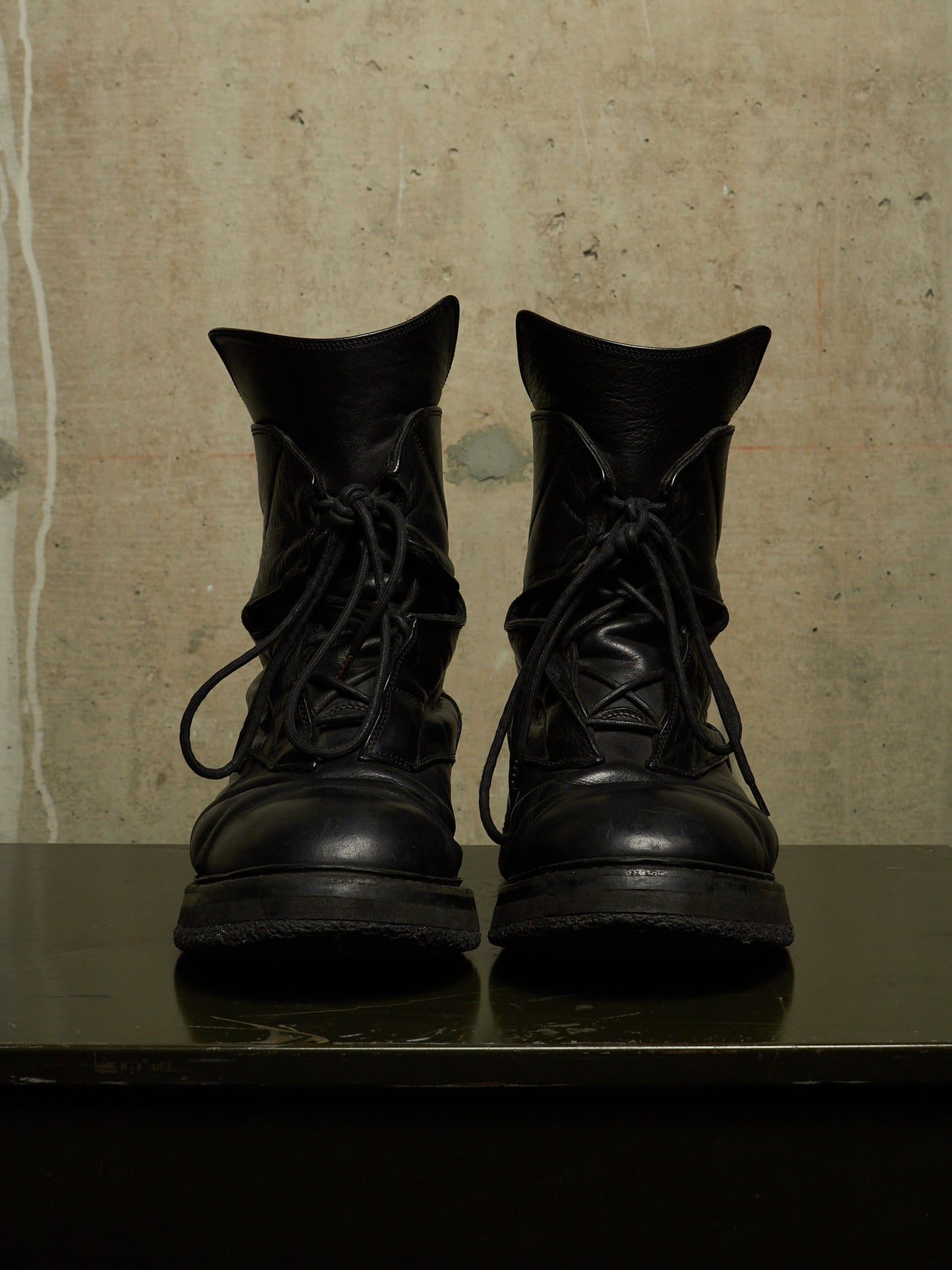 A pair of black leather lace-up combat boots with a rugged sole, displayed side-by-side on a dark reflective surface against a textured beige concrete wall.