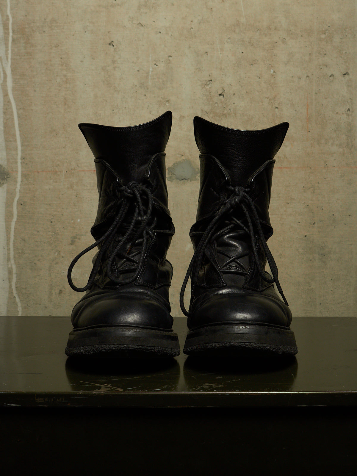 A pair of black leather lace-up combat boots with a rugged sole, displayed side-by-side on a dark reflective surface against a textured beige concrete wall.