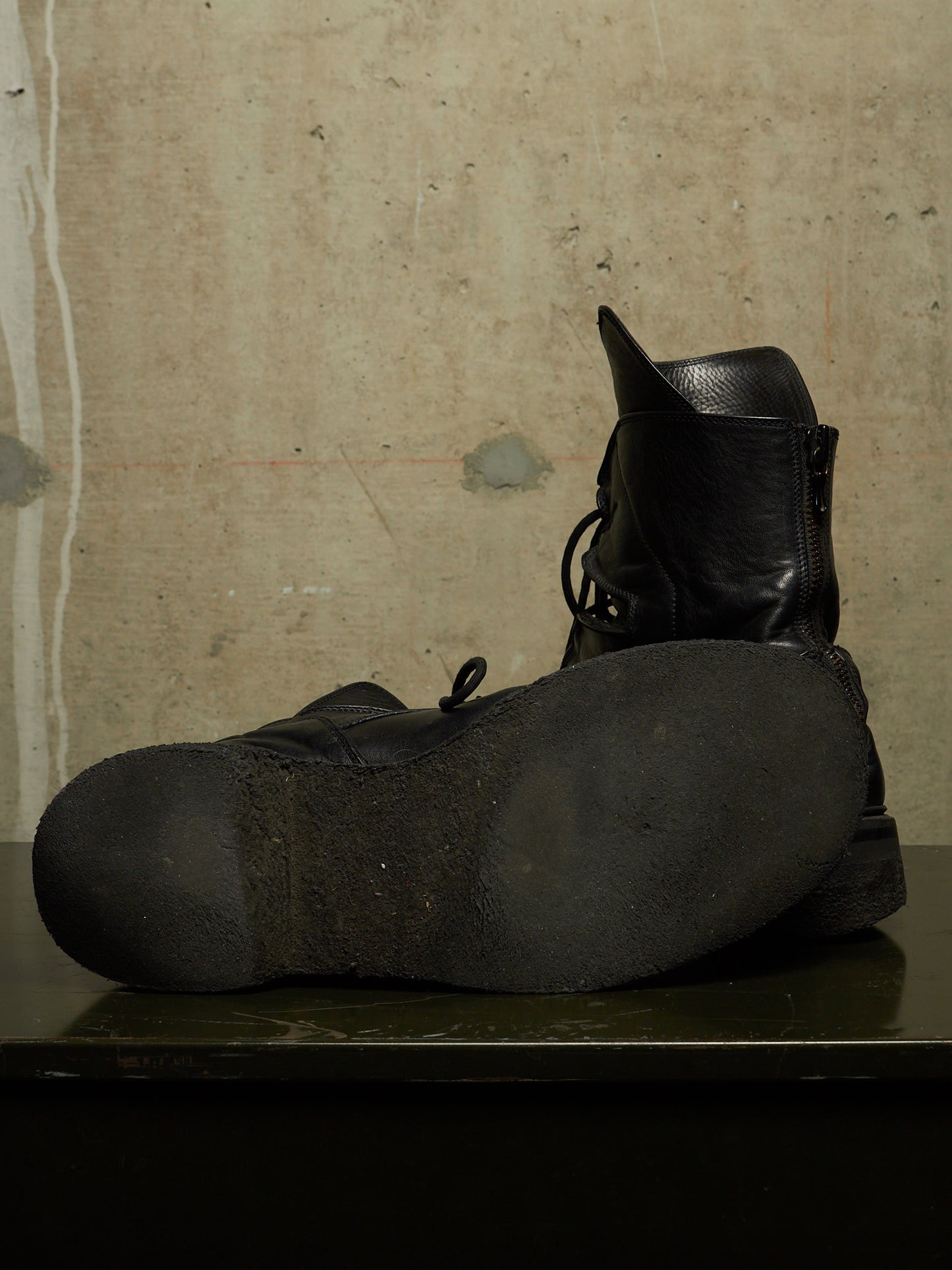 A pair of black leather lace-up boots, with one boot standing upright and the other lying on its side to show the sole, set against a textured concrete wall.