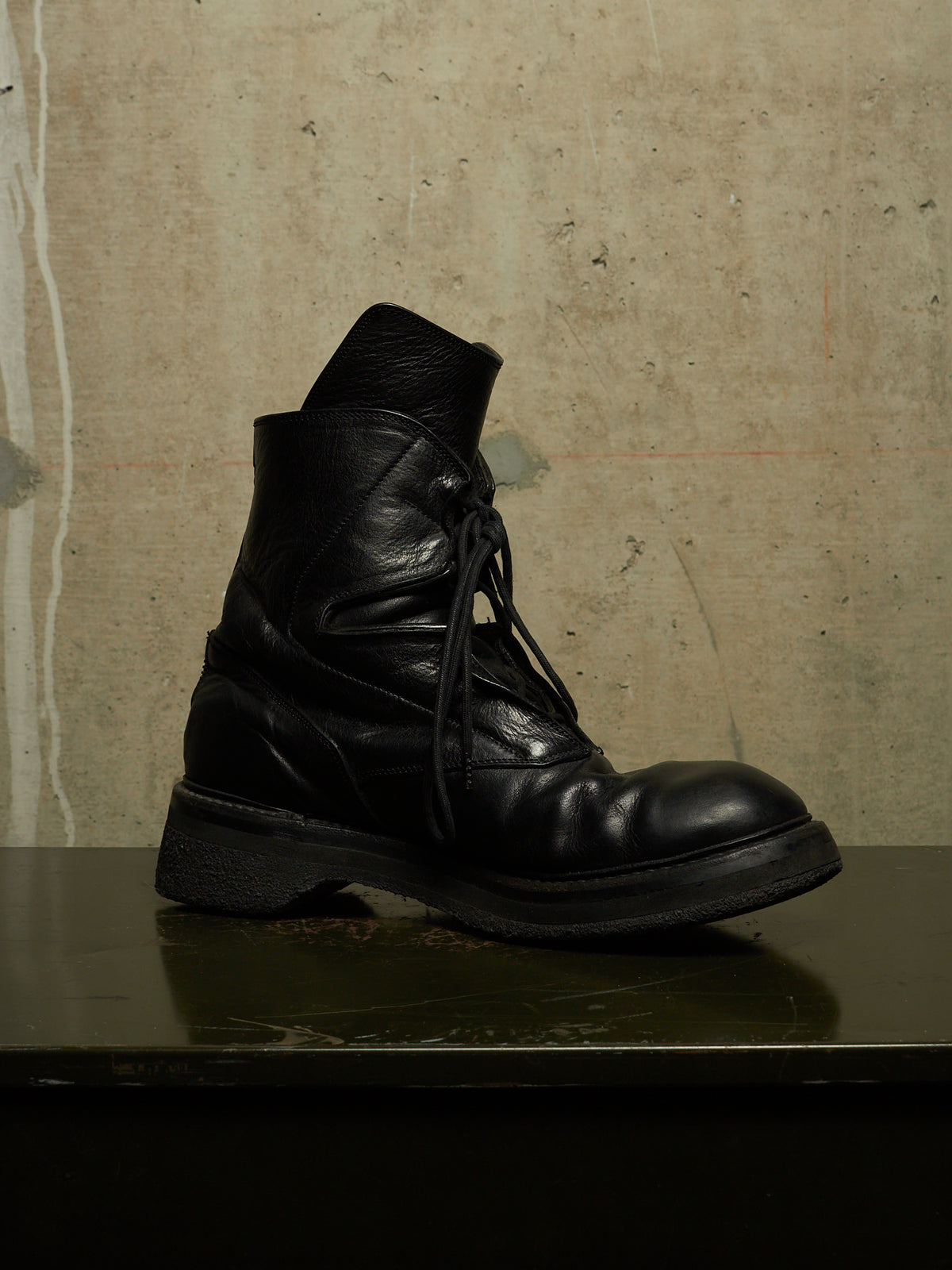 A single black leather lace-up boot with a chunky sole, photographed on a dark reflective surface against a textured beige wall.