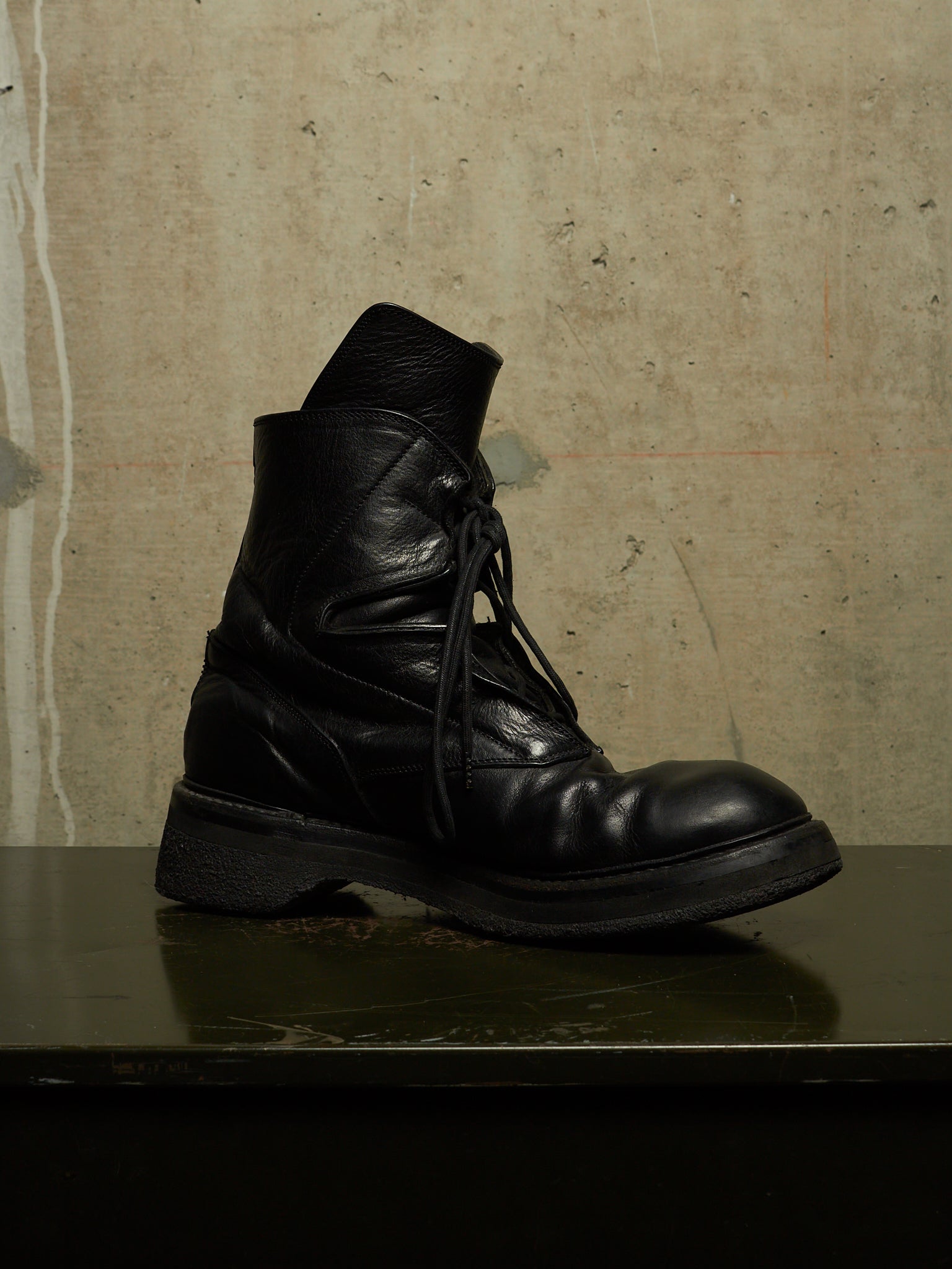 A single black leather lace-up boot with a chunky sole, photographed on a dark reflective surface against a textured beige wall.