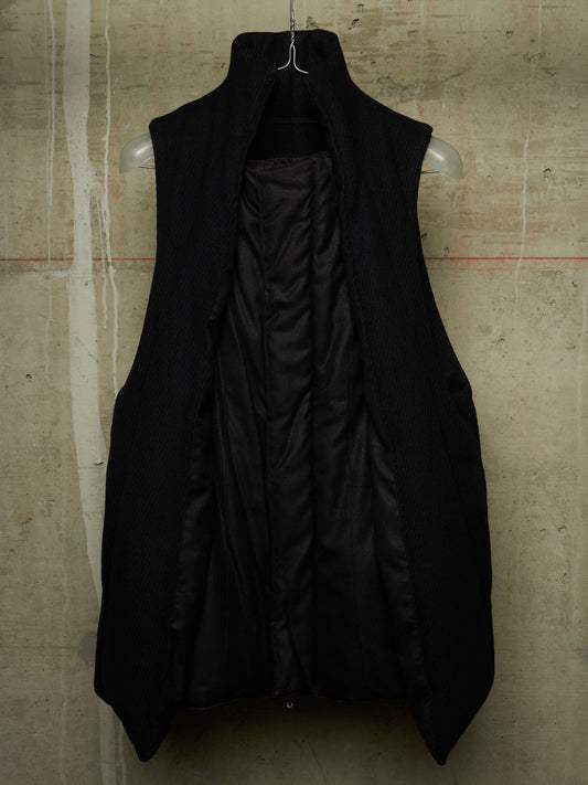 A black sleeveless coat with a high stand collar, hanging on a clear plastic hanger against a textured concrete wall.