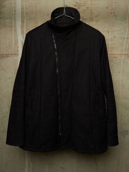 A black asymmetrical zip-up jacket with a high mock neck collar, hanging on a wire hanger against a textured concrete wall.