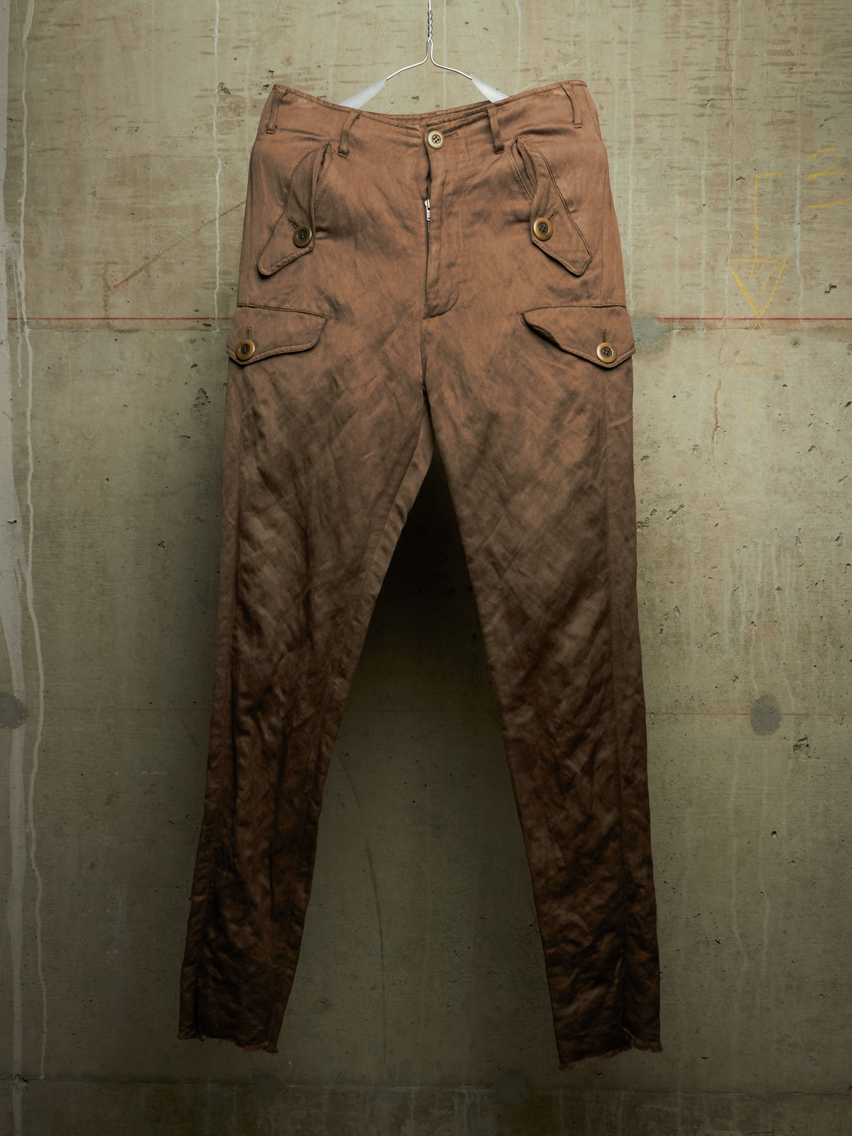 A pair of brown crinkled cargo pants hanging from a white plastic hanger against a textured concrete wall.