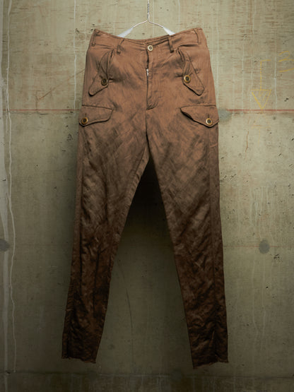 A pair of brown crinkled cargo pants hanging from a white plastic hanger against a textured concrete wall.