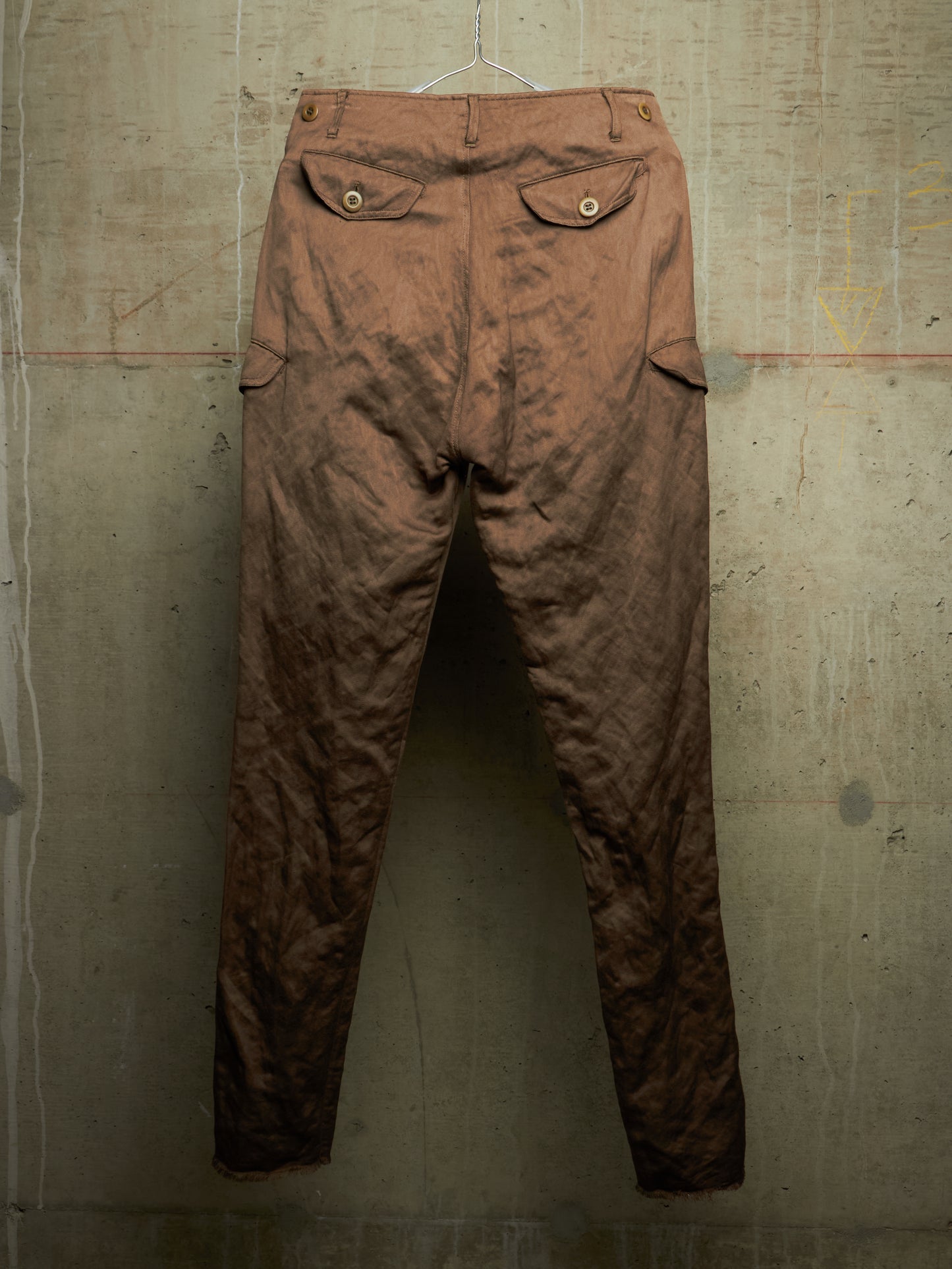 A pair of vintage brown trousers with a heavily distressed, faded, and crinkled texture, hanging against a textured concrete wall.