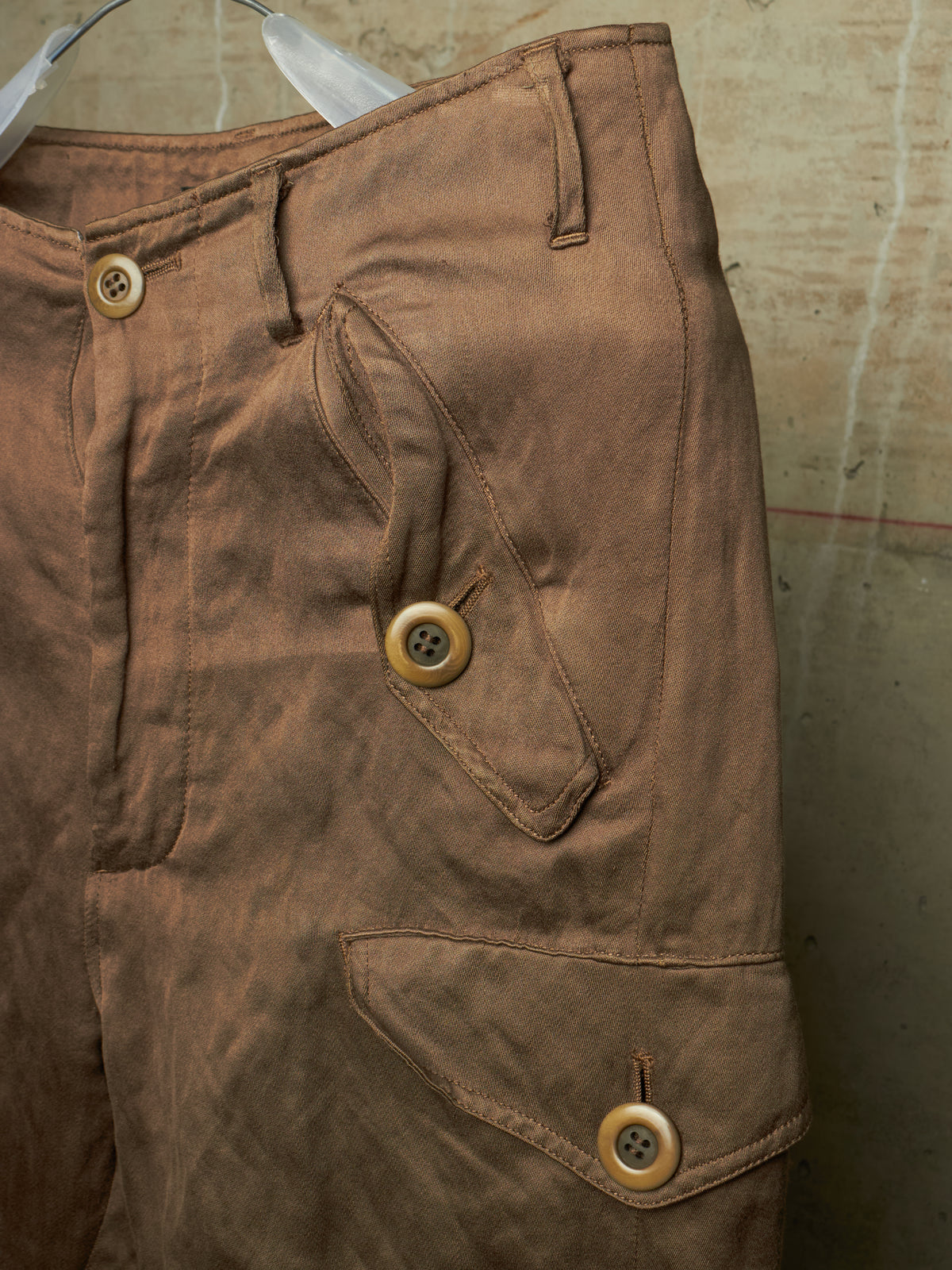 Close-up of brown canvas cargo pants hanging on a hanger, showing buttoned flap pockets and belt loops against a textured wall.
