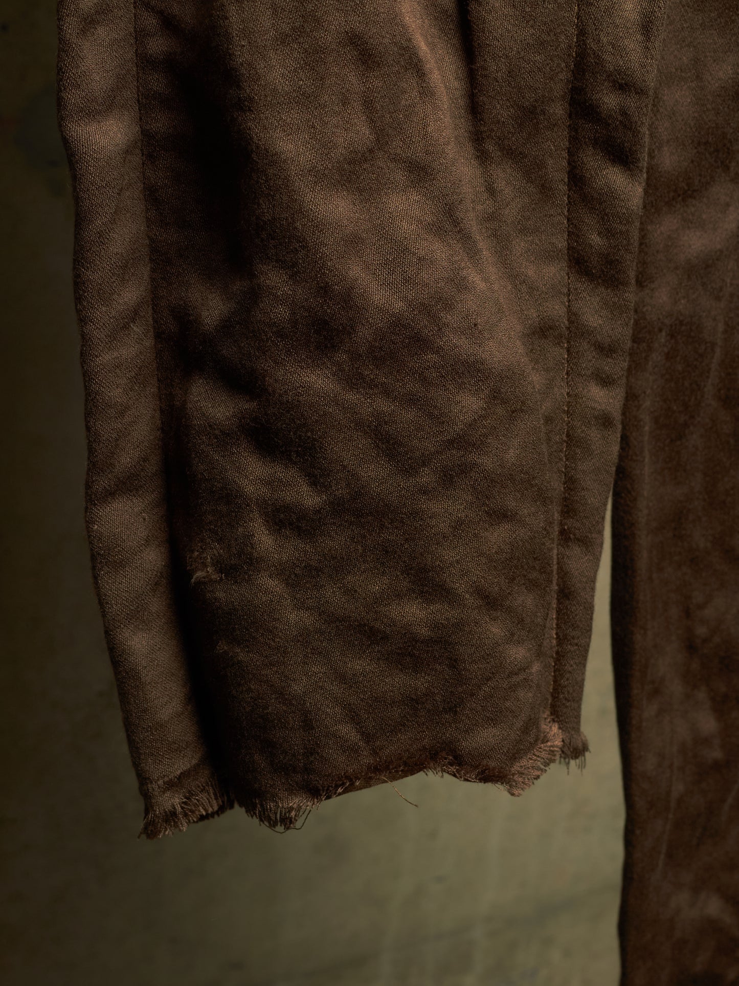 A close-up of a dark brown suede or leather fabric, showing its soft, textured surface and raw, frayed edges against a muted olive-gray background.