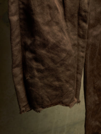 A close-up of a dark brown suede or leather fabric, showing its soft, textured surface and raw, frayed edges against a muted olive-gray background.