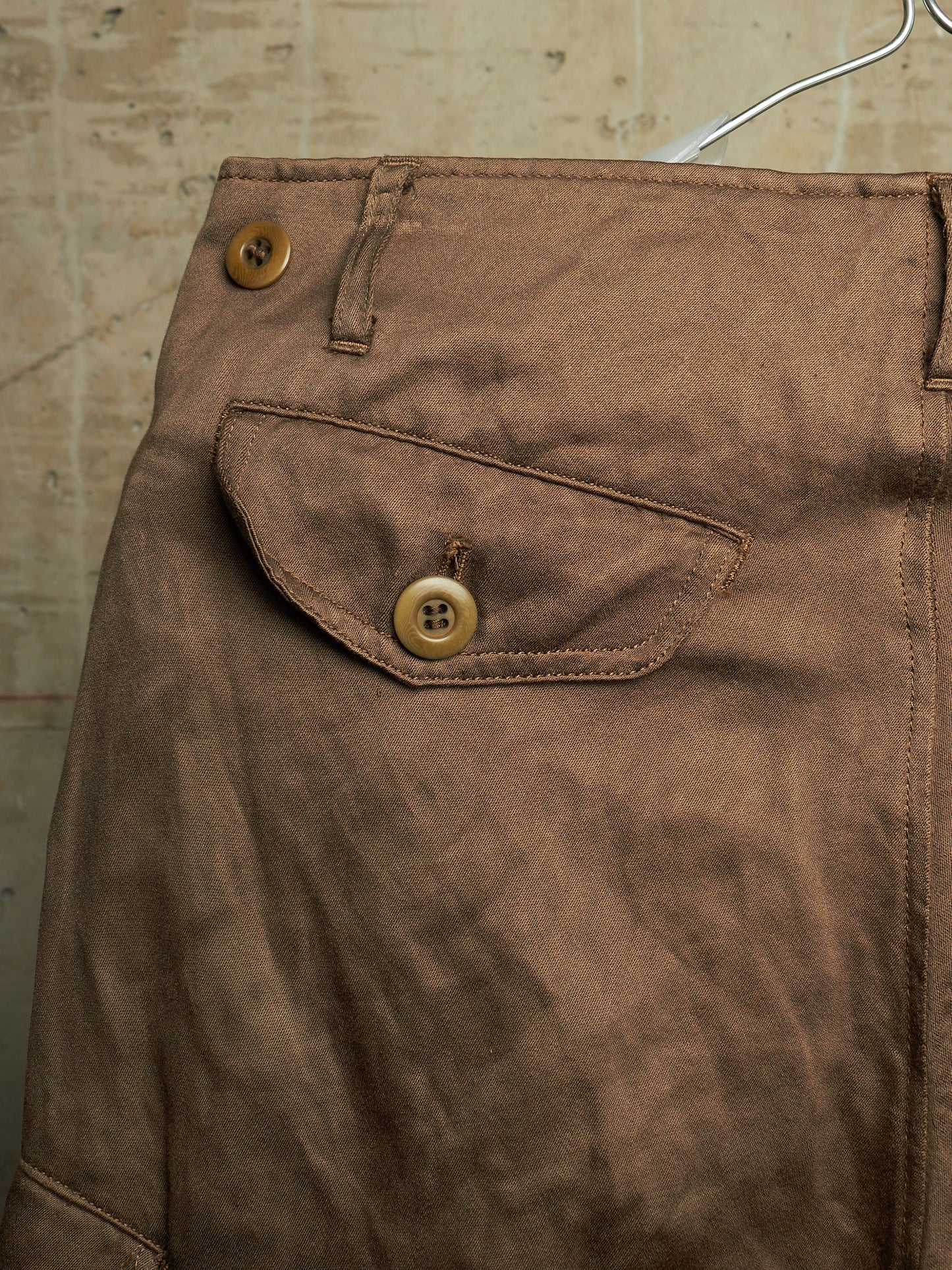 Close-up of brown fabric clothing hanging on a hanger, showing a buttoned flap pocket and belt loops against a textured beige wall.