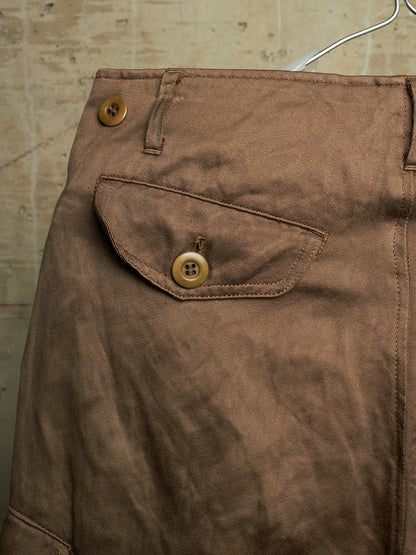 Close-up of brown fabric clothing hanging on a hanger, showing a buttoned flap pocket and belt loops against a textured beige wall.
