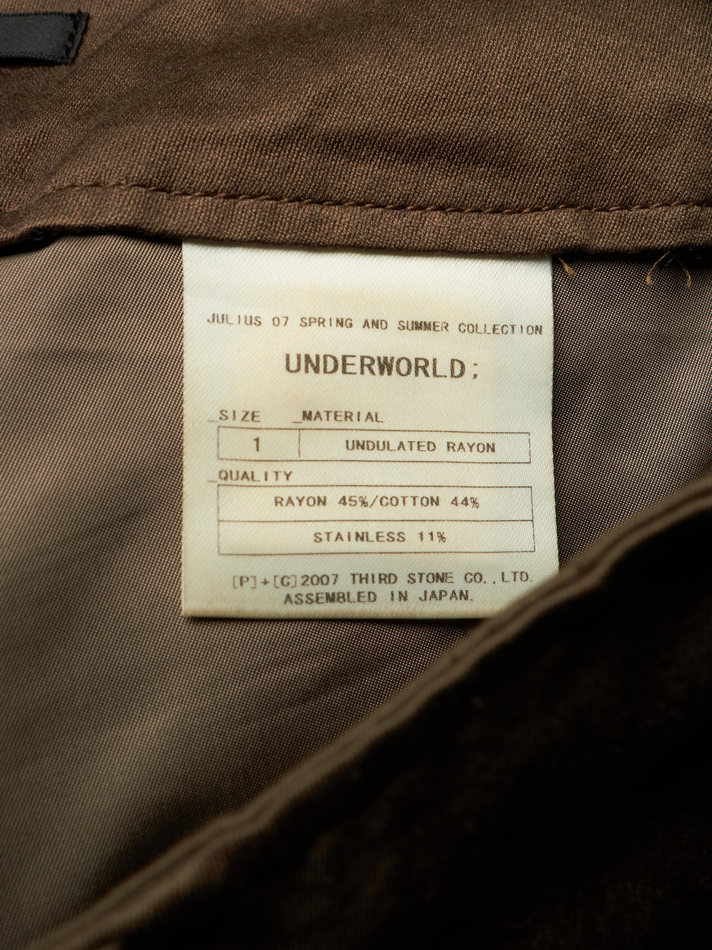 Close-up of the inside collar of a brown garment showing a white paper size and material label that reads 'UNDERWORLD' and 'Julius 07 Spring and Summer Collection'.