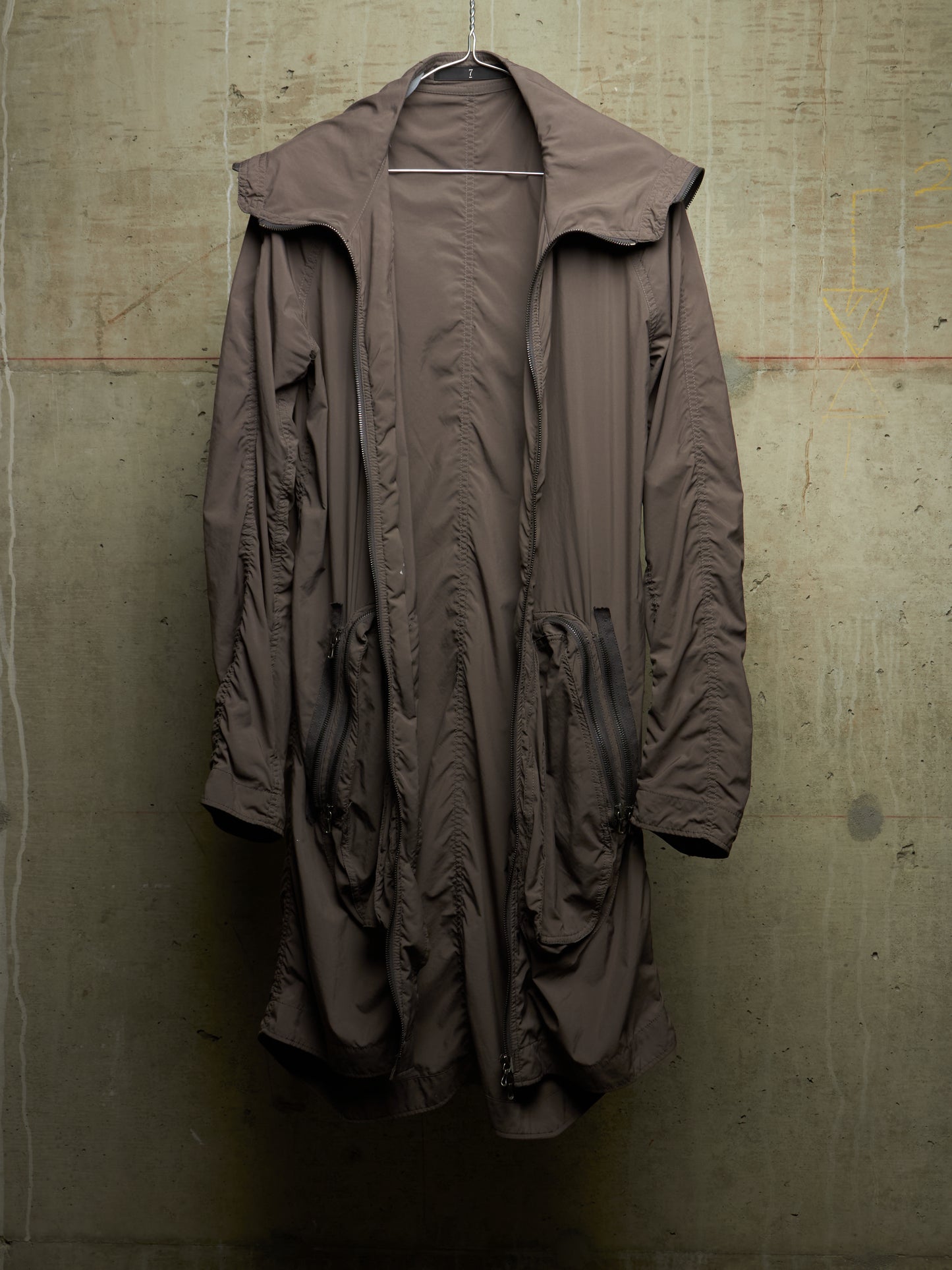 A long, oversized brown hooded jacket with a high collar and front zipper, hanging against a textured concrete wall.