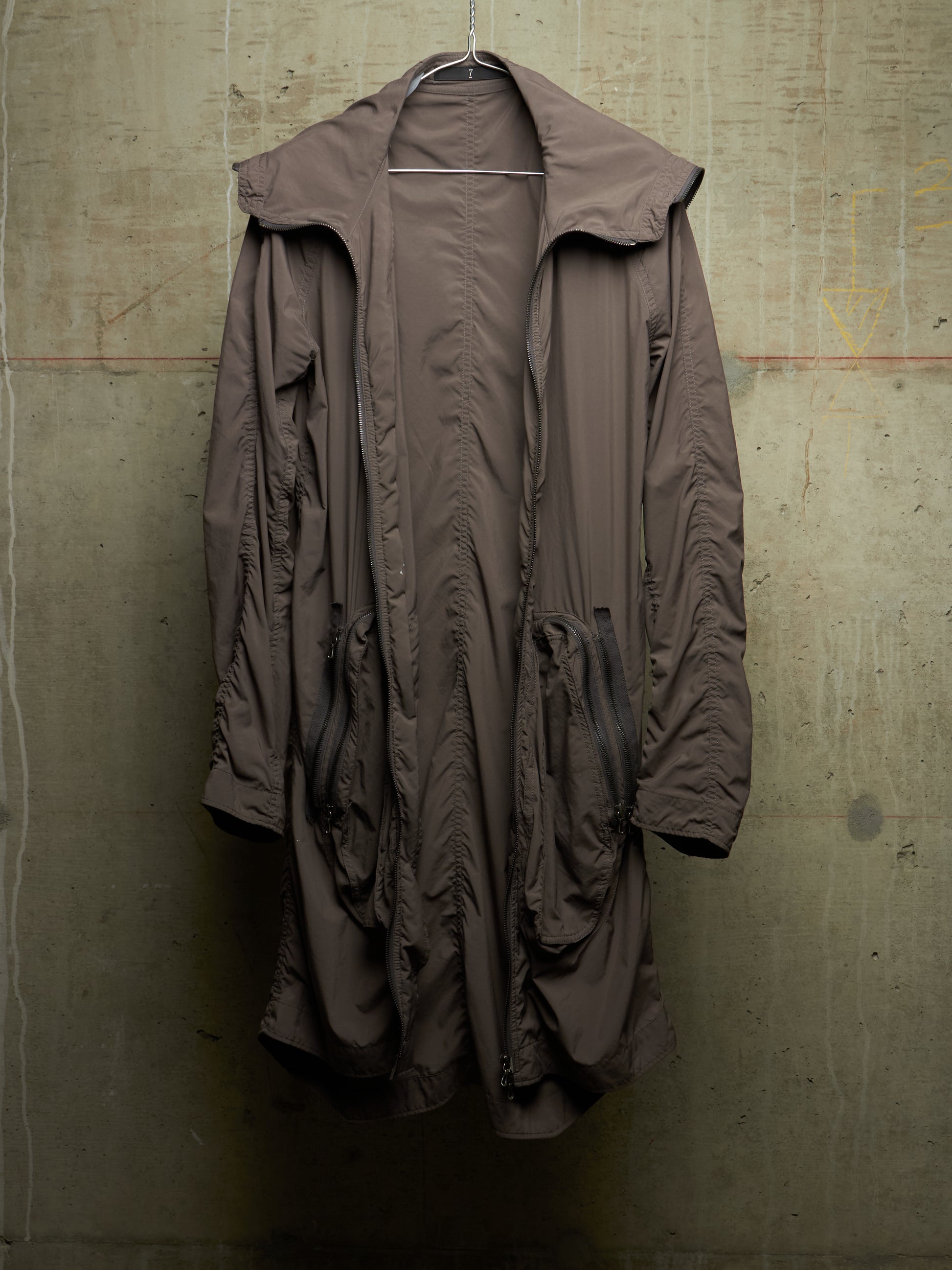 A long, oversized brown hooded jacket with a high collar and front zipper, hanging against a textured concrete wall.