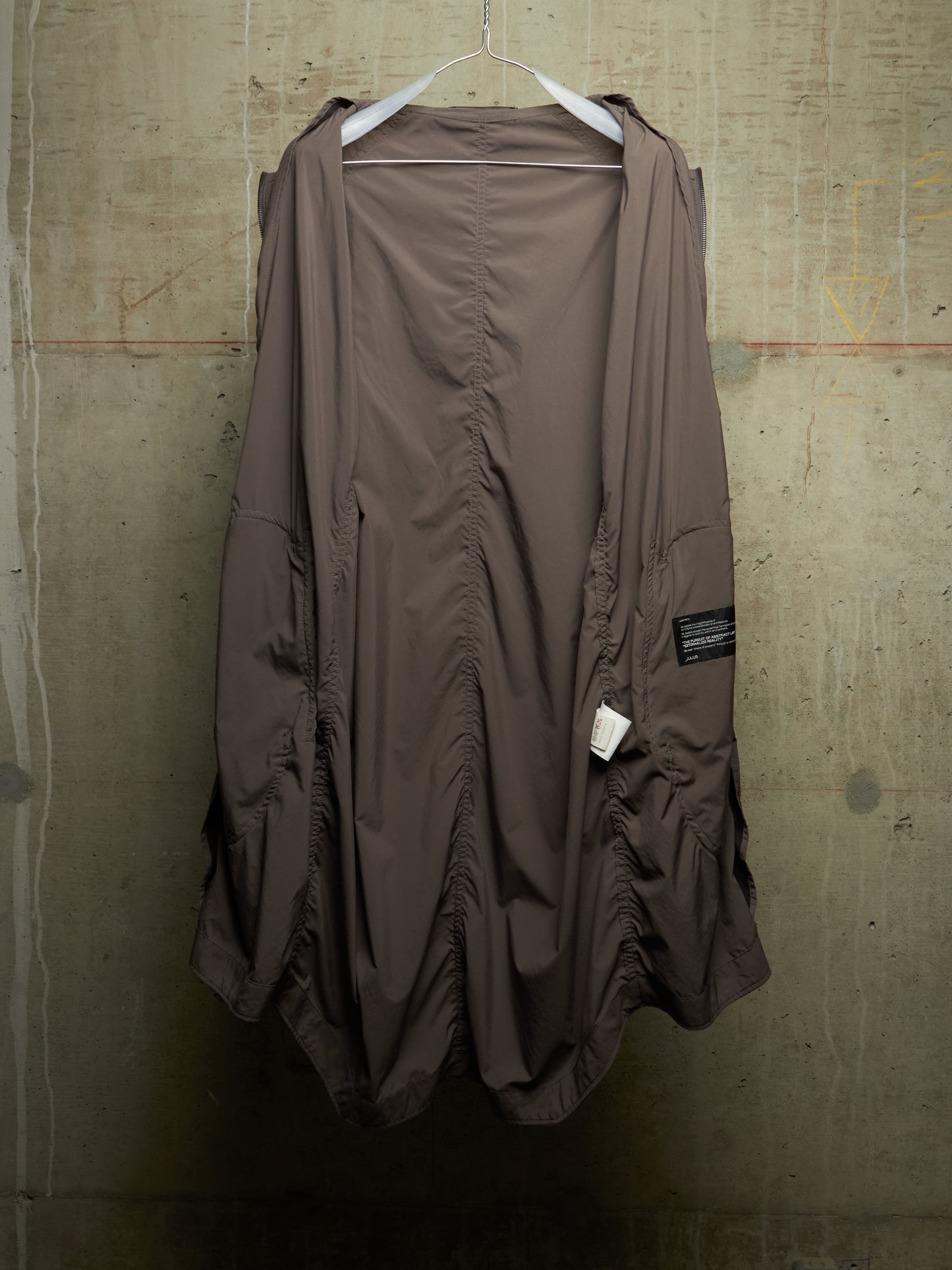 A brown, loose-fitting, draped jacket or cover-up with ruched details, hanging on a white hanger against a textured concrete wall.