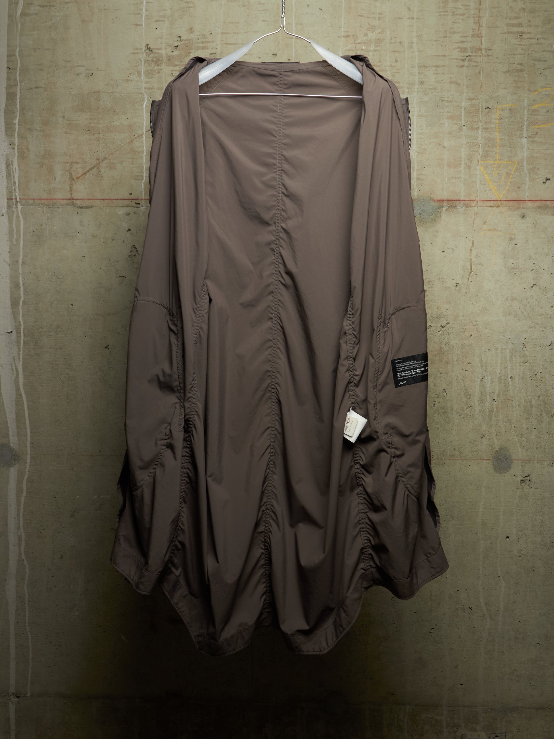 A brown, loose-fitting, draped jacket or cover-up with ruched details, hanging on a white hanger against a textured concrete wall.