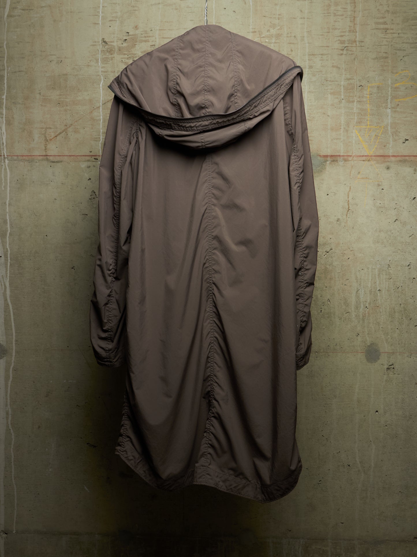 A long, draped hooded coat in a muted taupe-brown color, hanging against a textured concrete wall.