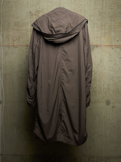 A long, draped hooded coat in a muted taupe-brown color, hanging against a textured concrete wall.