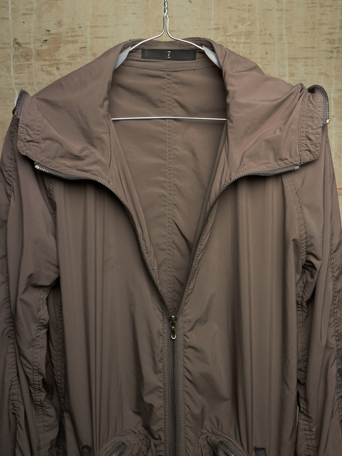 A brown lightweight hooded jacket hanging on a metal hanger against a textured beige wall.