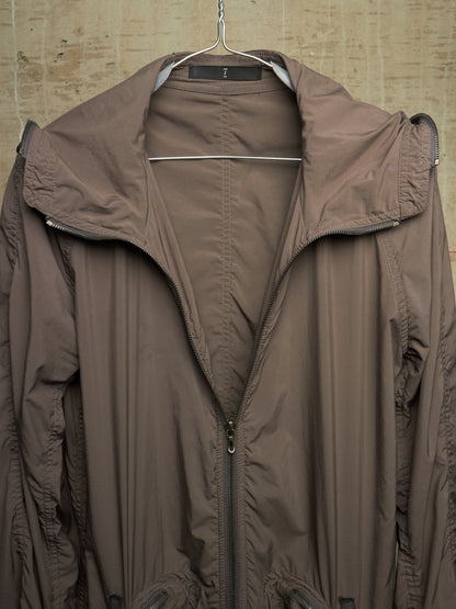 A brown lightweight hooded jacket hanging on a metal hanger against a textured beige wall.