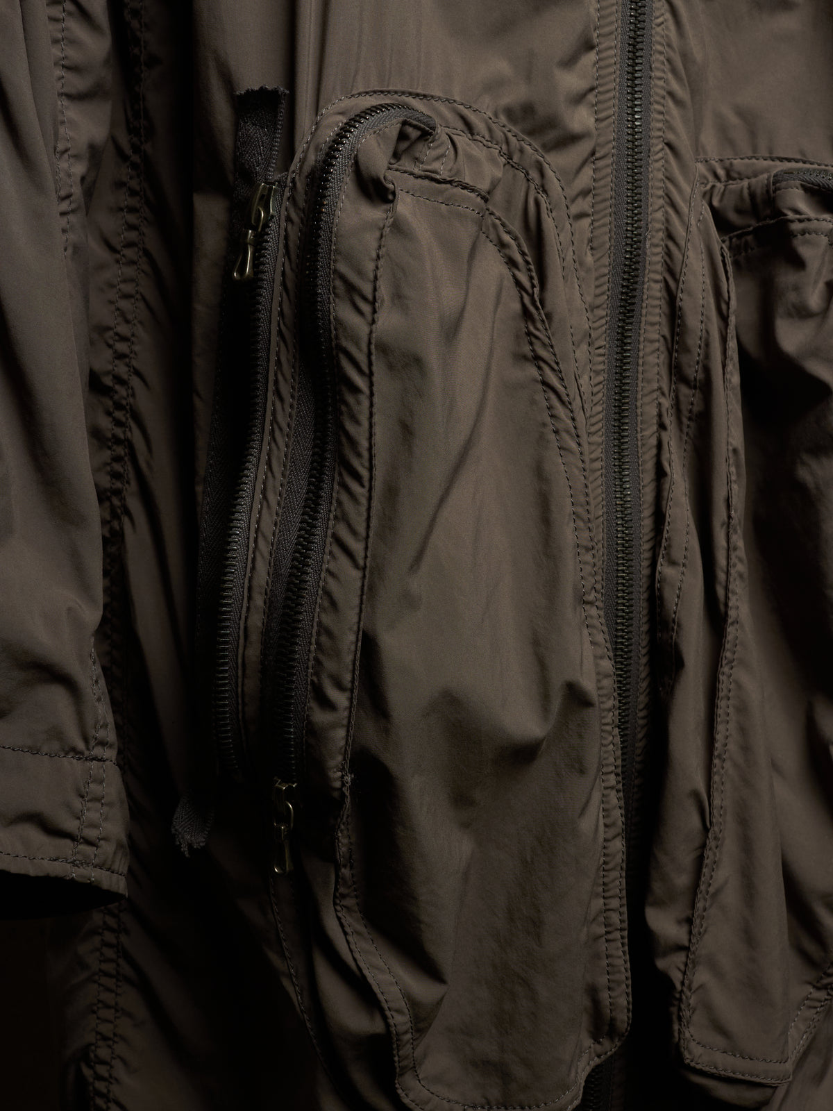 Close-up of a dark olive-brown technical jacket with multiple zippered pockets and stitched panel details.