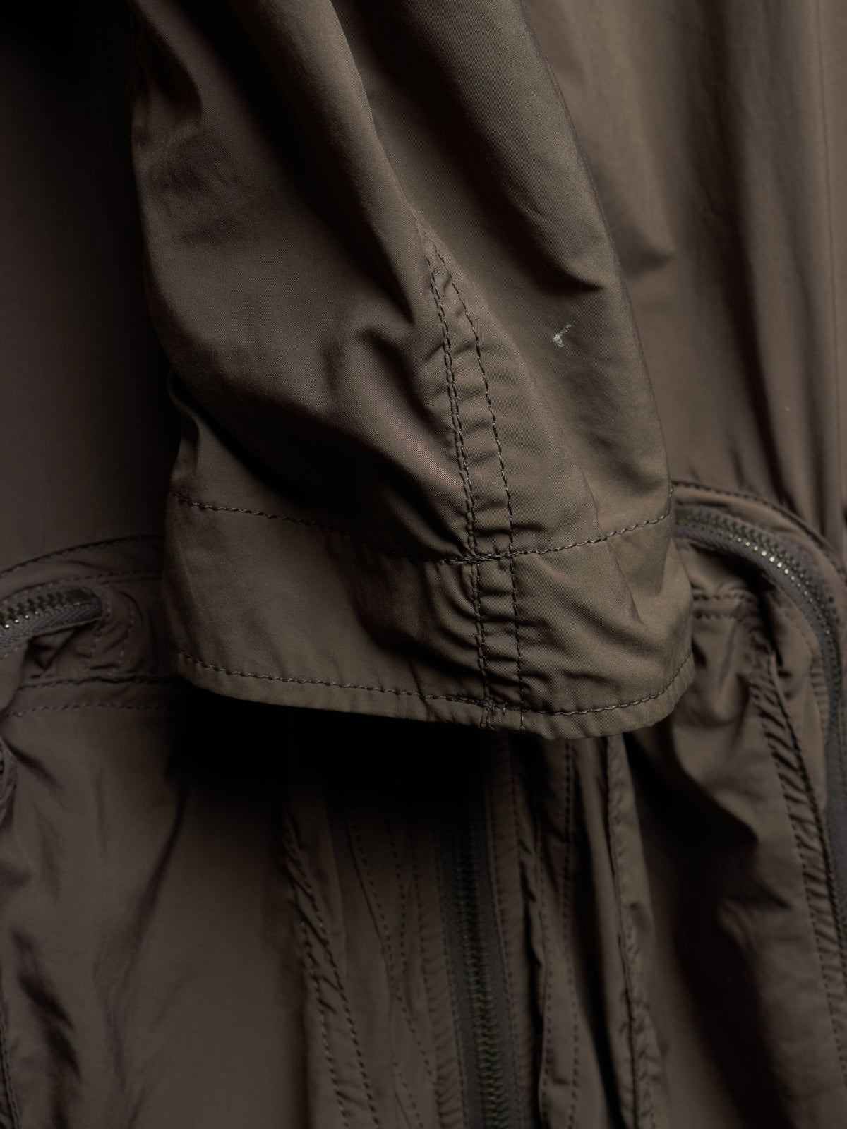 Close-up detail of a dark brown waterproof jacket, showing the sleeve cuff with precise stitching, a drawcord, and a zipper pocket.