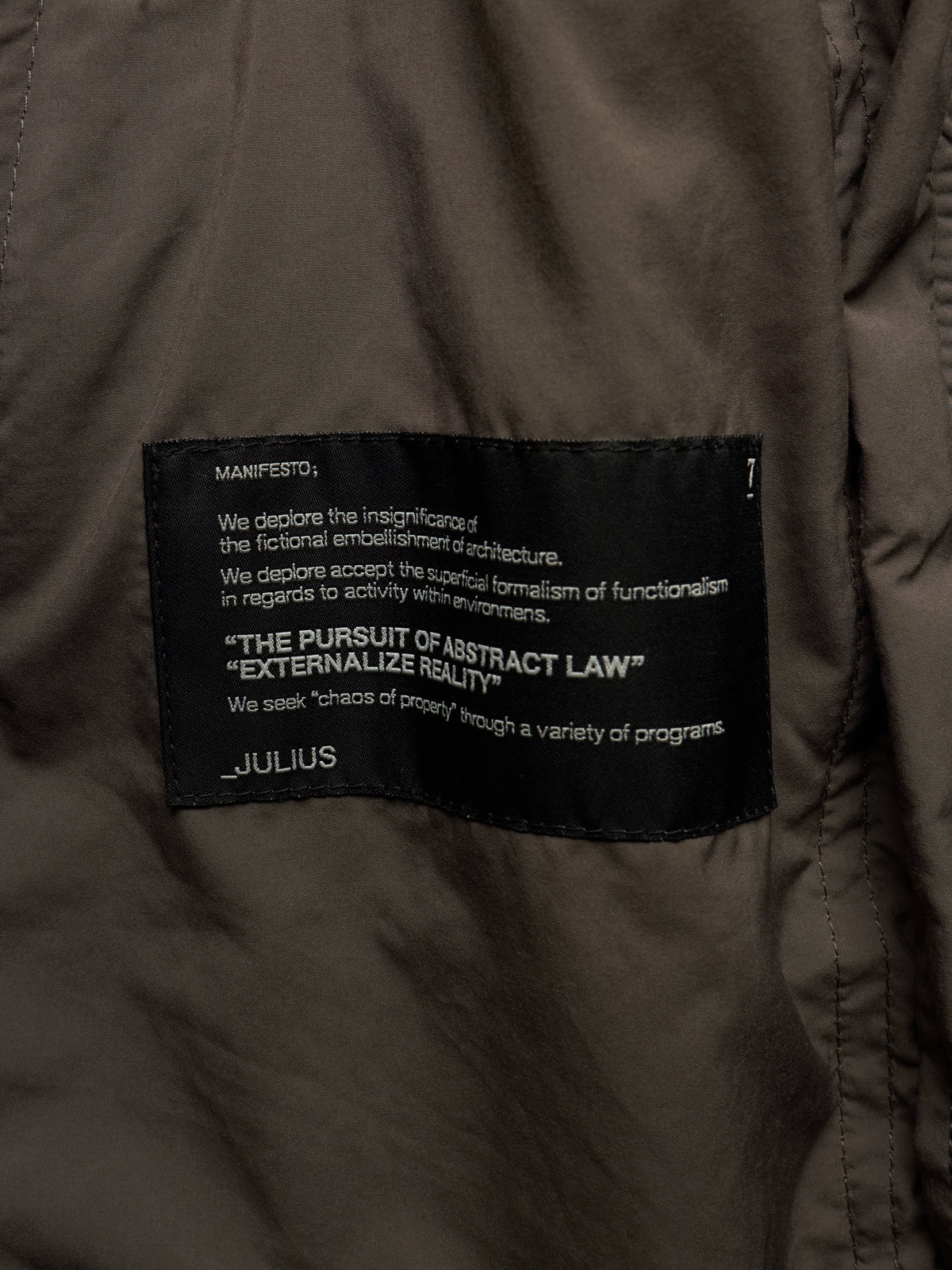 Close-up of a dark olive brown puffer jacket showing a black woven label with white text that reads 'MANIFESTO' and 'JULIUS'.