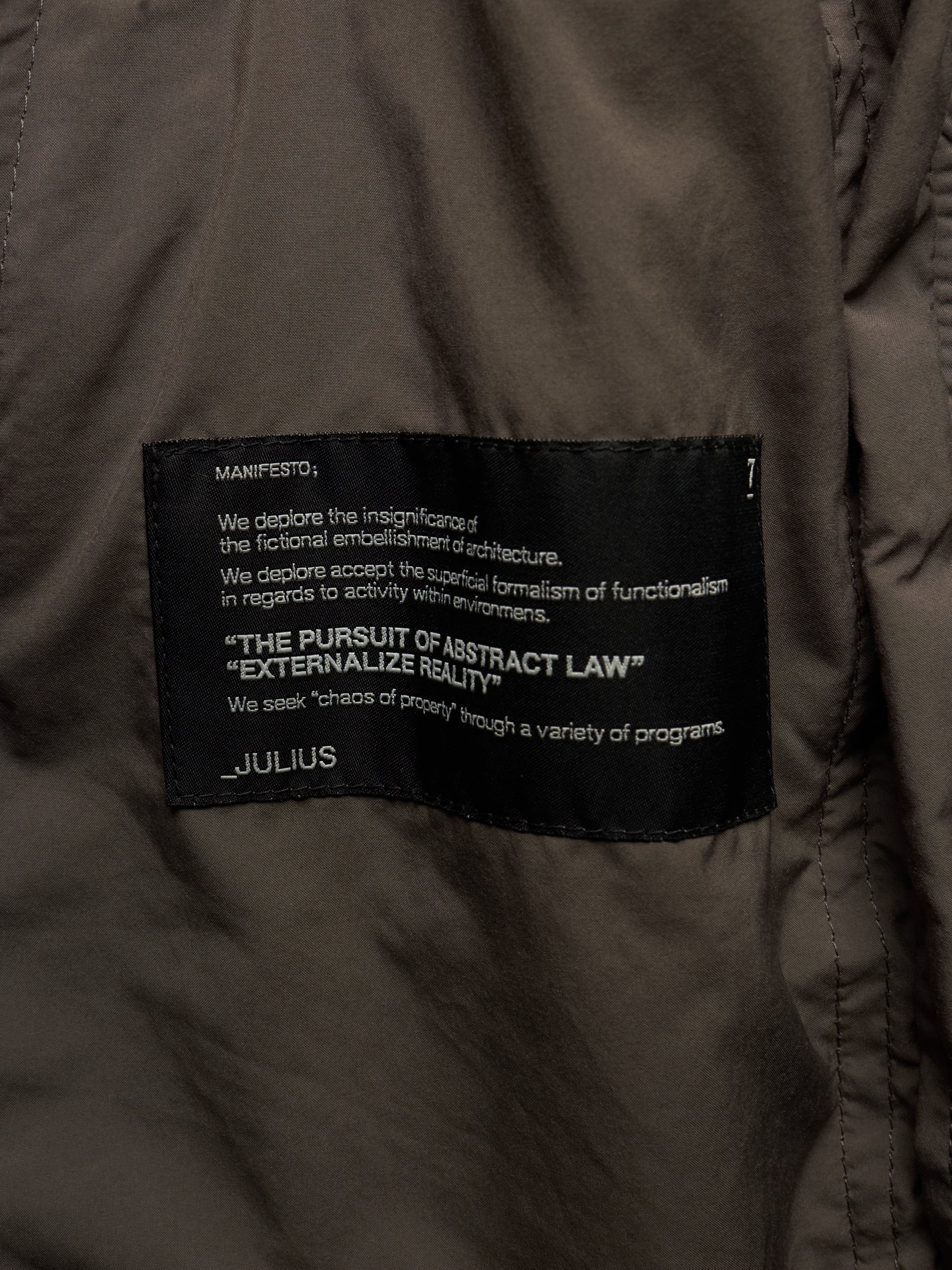 Close-up of a dark olive brown puffer jacket showing a black woven label with white text that reads 'MANIFESTO' and 'JULIUS'.
