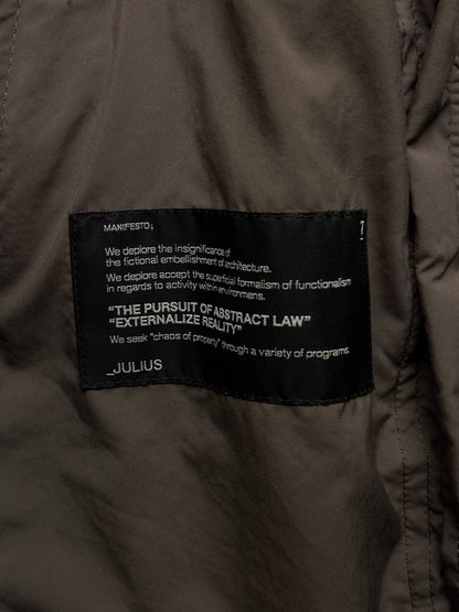 Close-up of a dark olive brown puffer jacket showing a black woven label with white text that reads 'MANIFESTO' and 'JULIUS'.