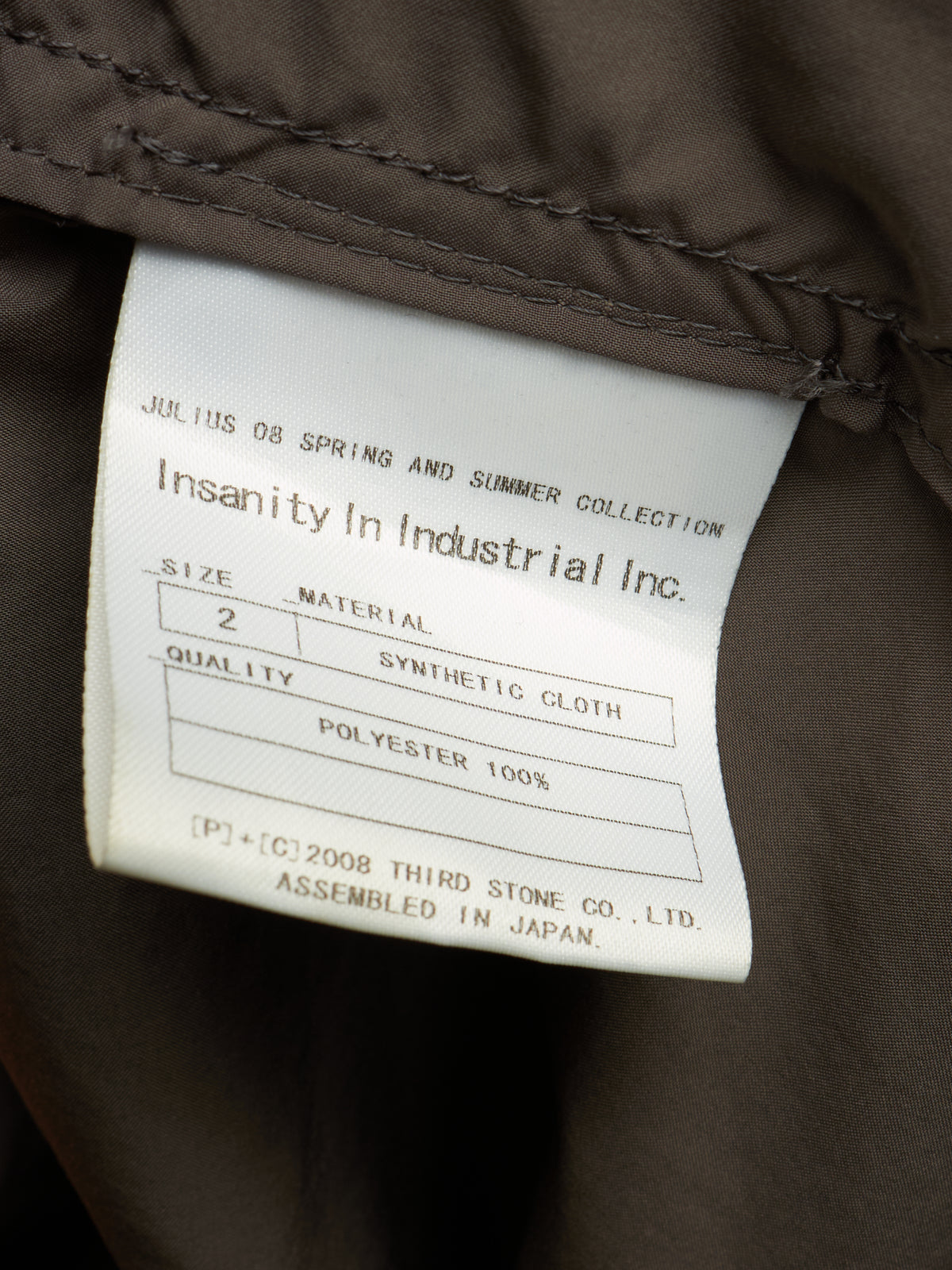 Close-up of a garment's interior label reading 'Insanity In Industrial Inc.' with size, material, and polyester details, attached to dark brown fabric.