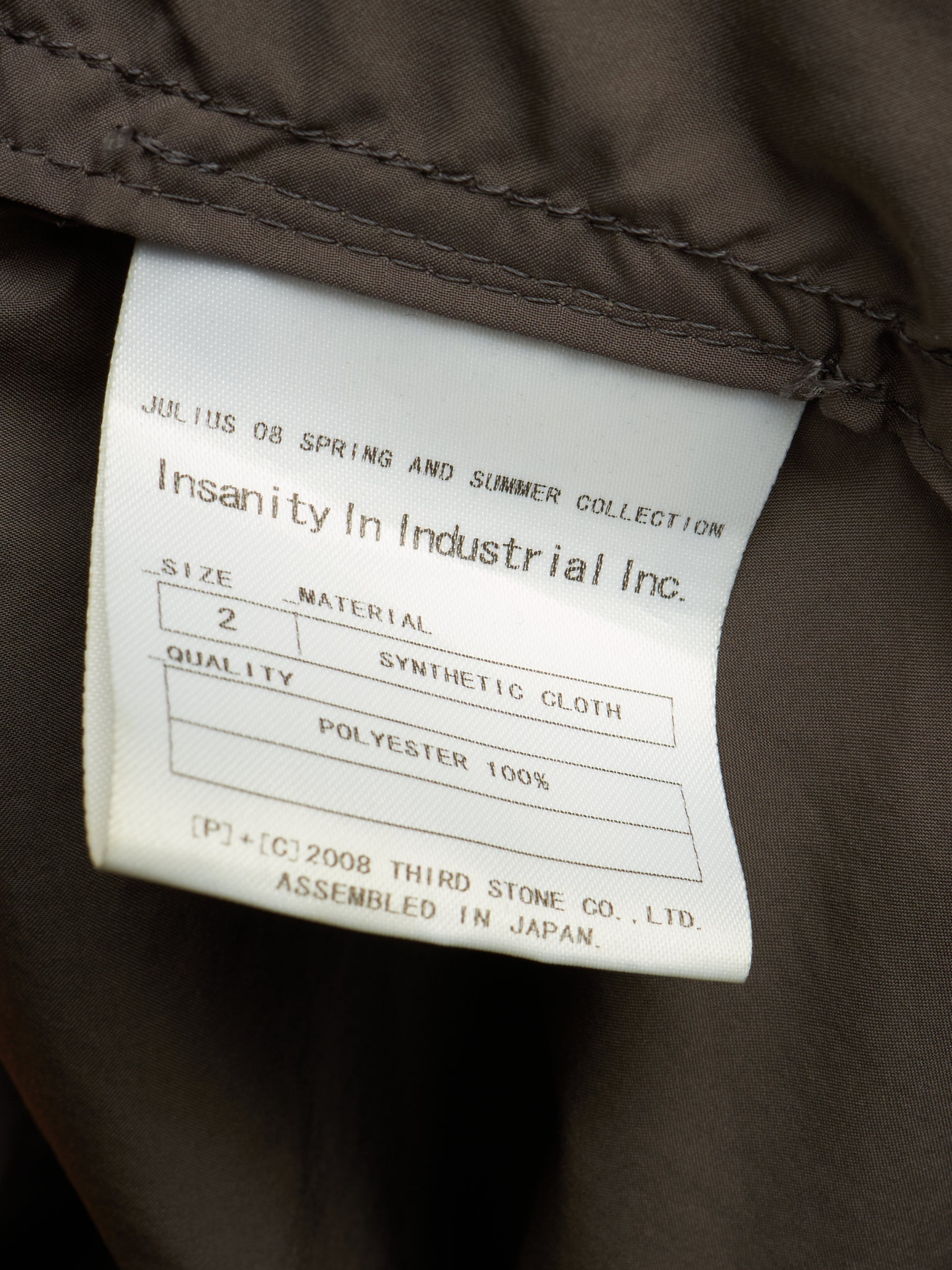 Close-up of a garment's interior label reading 'Insanity In Industrial Inc.' with size, material, and polyester details, attached to dark brown fabric.