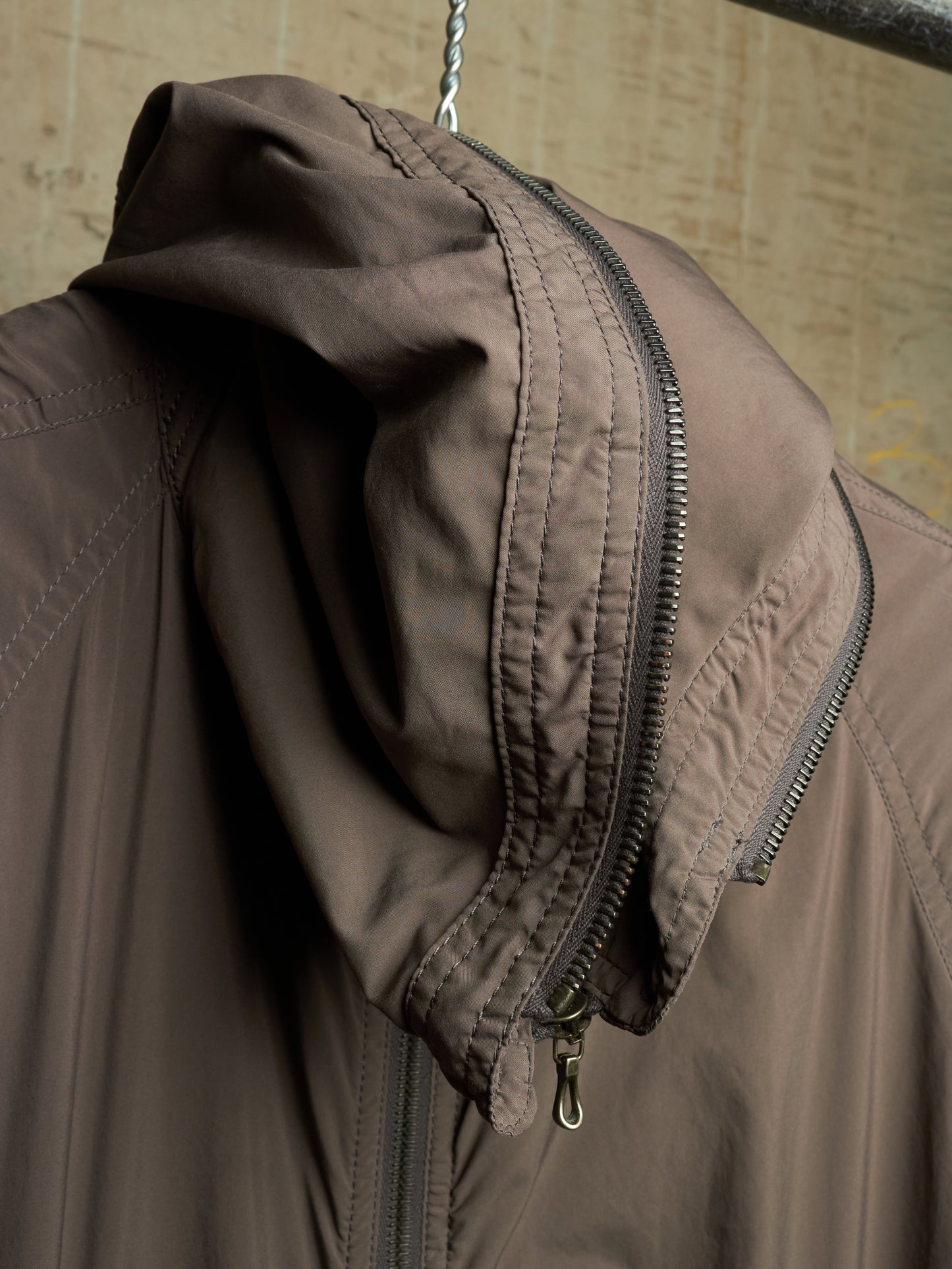 Close-up of a brown hooded jacket hanging on a metal hanger, showing the hood lining, zipper details, and textured fabric against a neutral beige background.