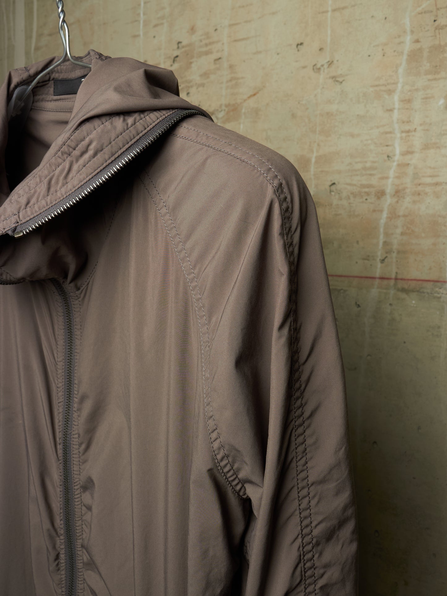A taupe-brown hooded jacket hanging on a metal hanger against a textured concrete wall.