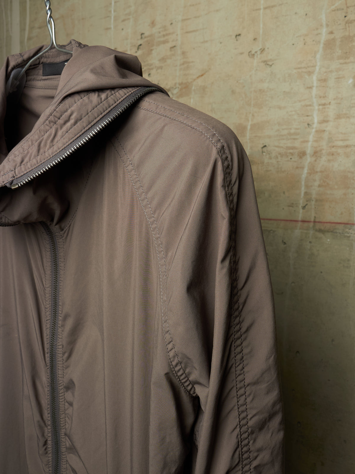 A taupe-brown hooded jacket hanging on a metal hanger against a textured concrete wall.