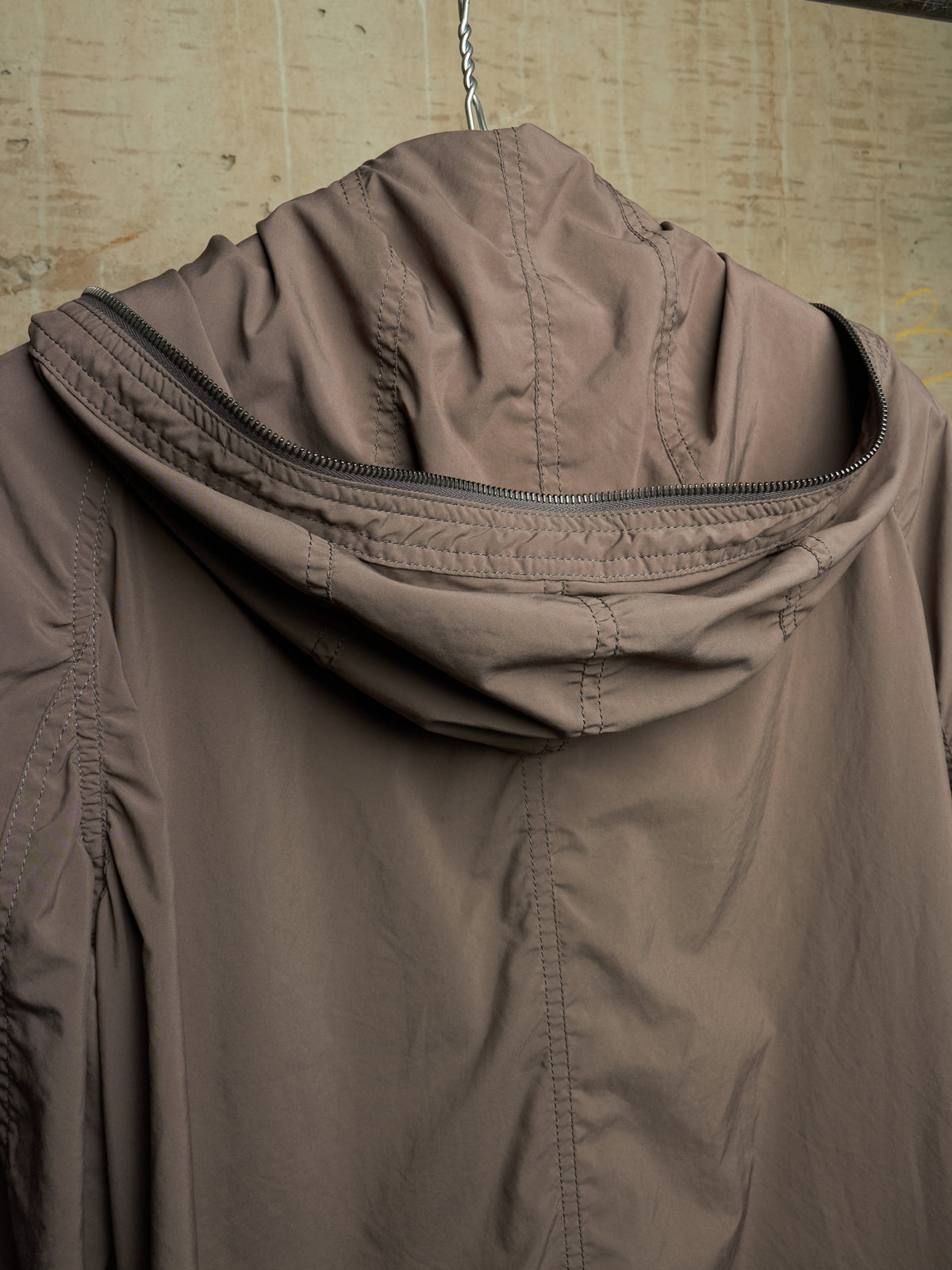 A close-up of a brown hooded jacket hanging against a textured beige wall, showing the back of the hood and upper back panel.