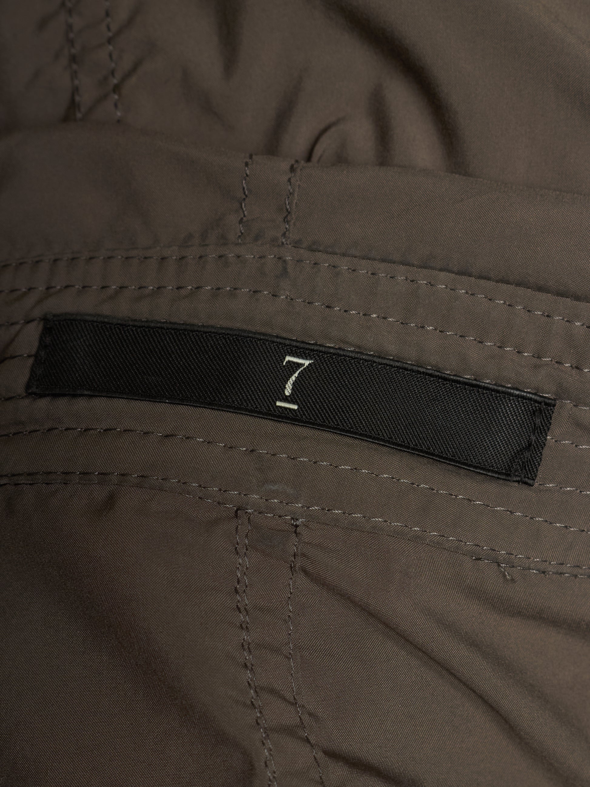 Close-up of a dark brown or taupe fabric garment with a black woven label featuring a white stylized number 7.