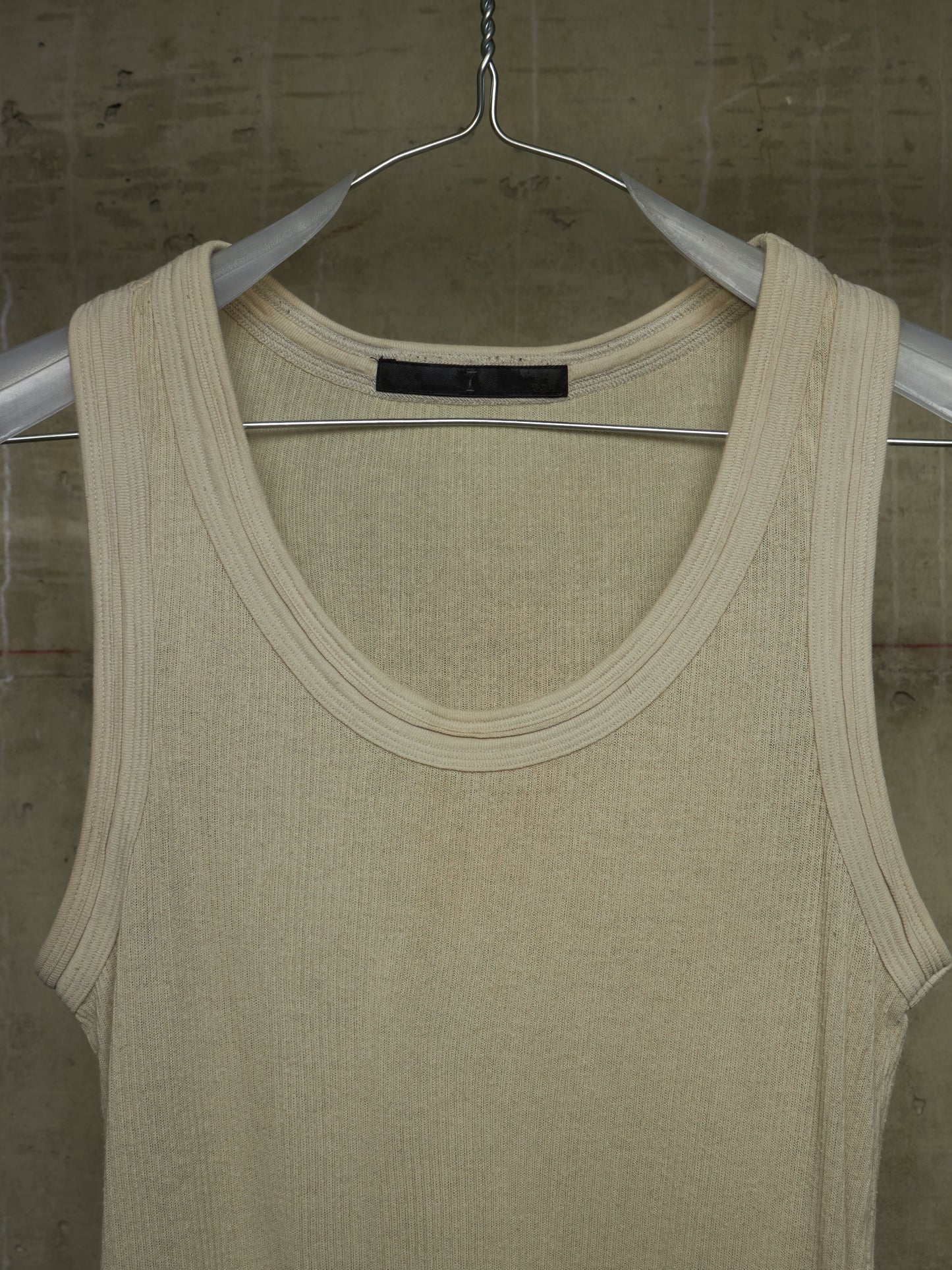 A cream-colored ribbed knit tank top hanging on a silver metal hanger against a textured concrete wall.